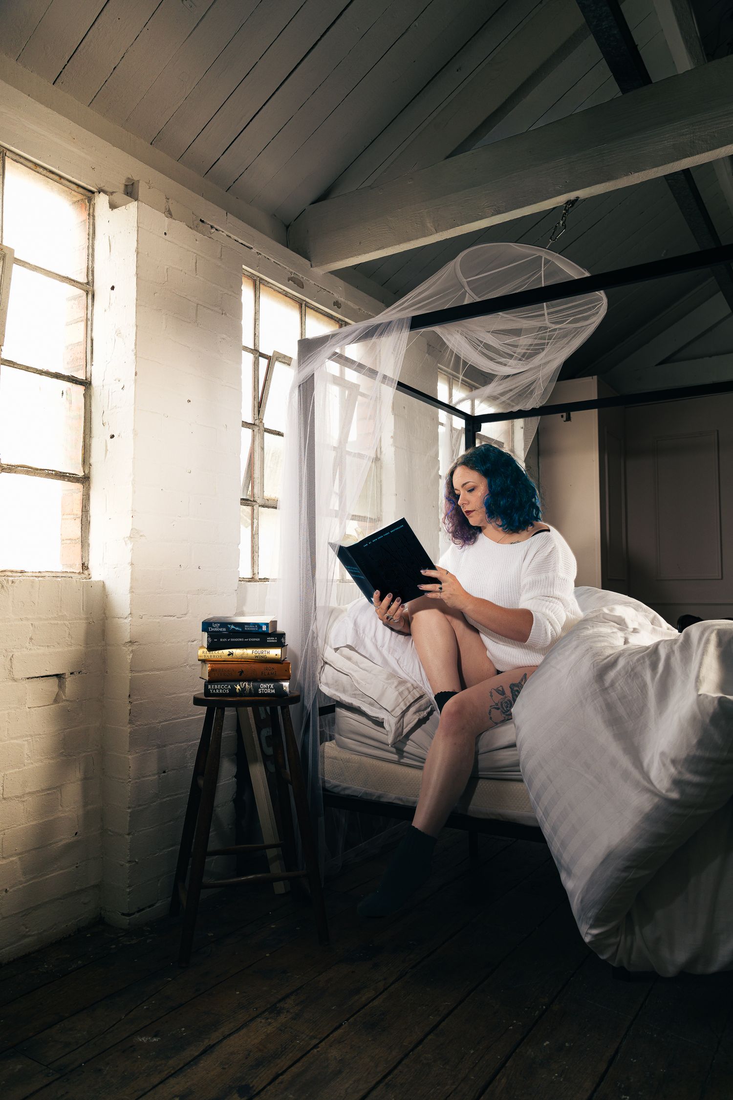 Person reading in bed surrounded by white linens in a rustic room with windows.