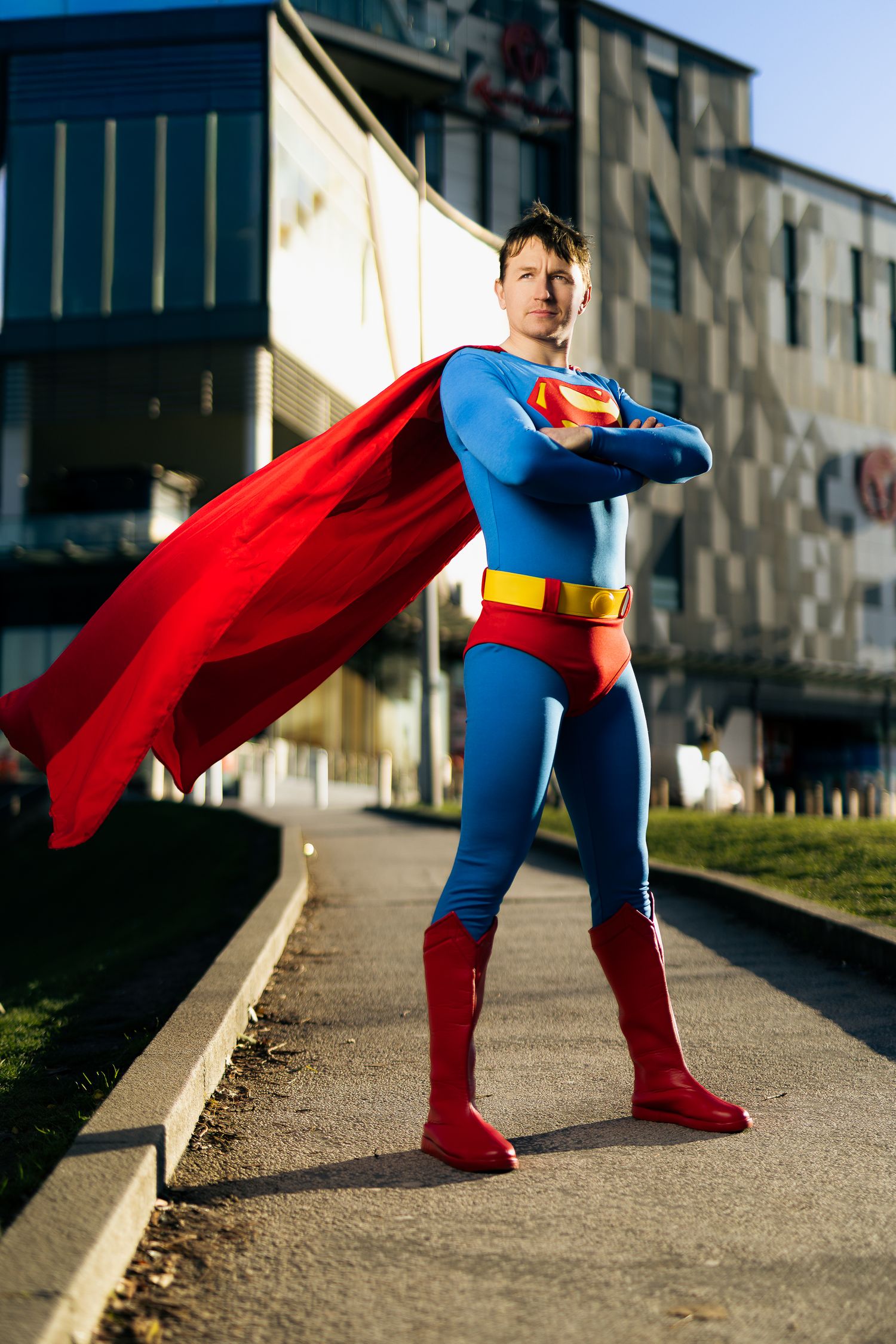 Superman cosplay standing with arms crossed, red cape flowing, outside a modern building in warm sunlight.