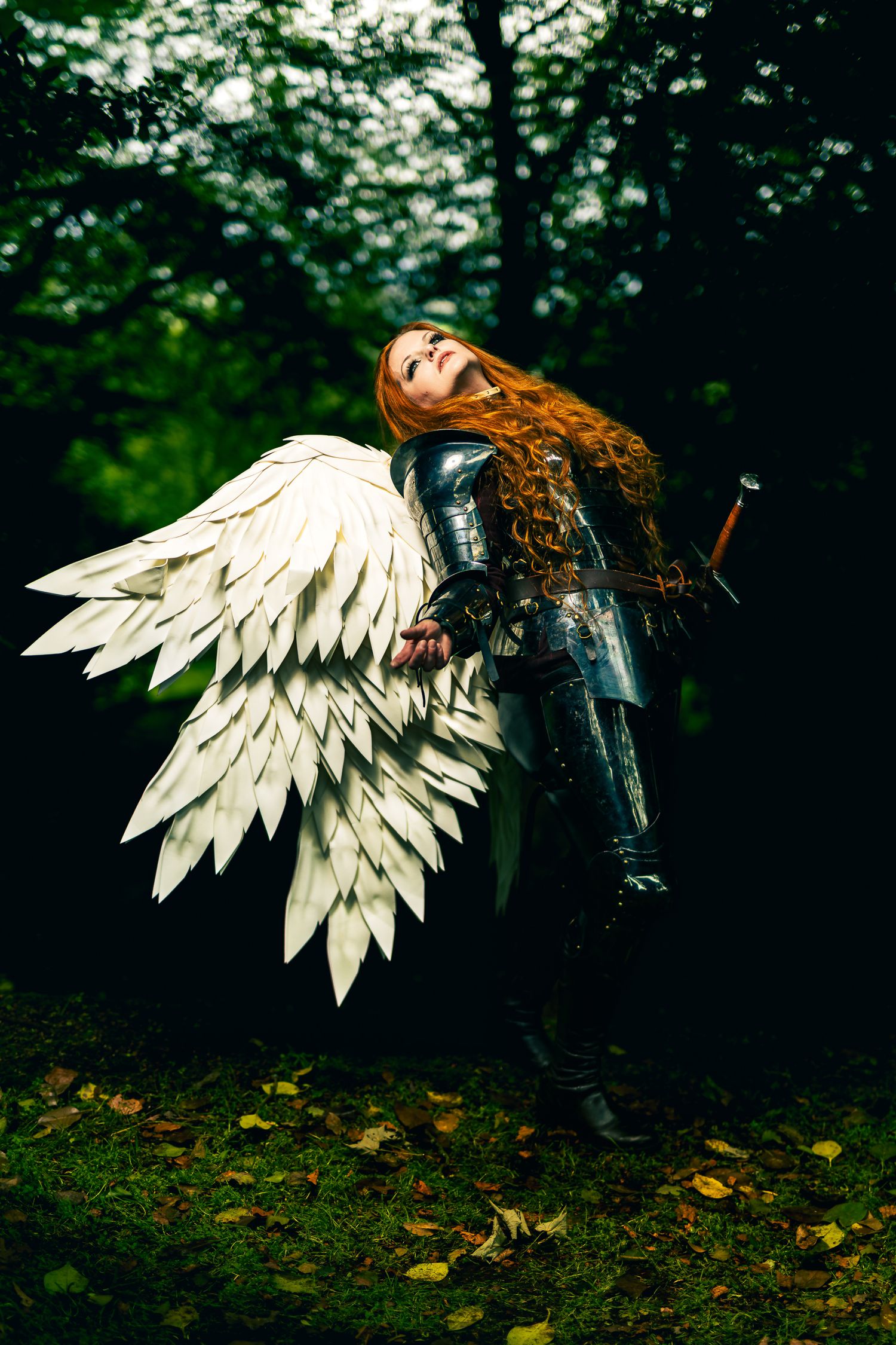 A red-haired woman in dark armor with large white angel wings poses dramatically in a forest setting.