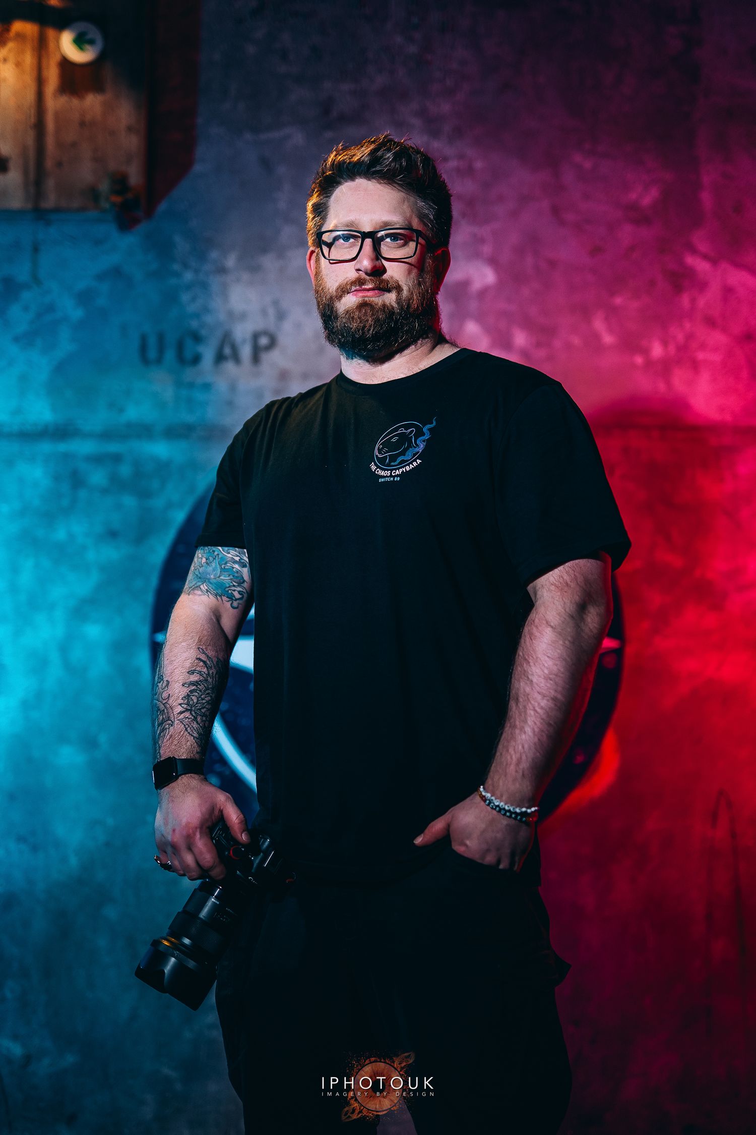 Bearded man with glasses holding a camera, wearing a black t-shirt, with blue and red lighting backdrop.