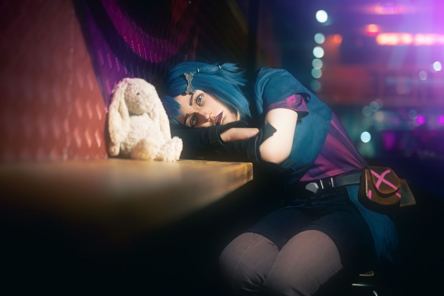 Blue-haired Jinx/powder cosplayer resting on a bar beside a plush bunny, lit by pink and blue neon bokeh.