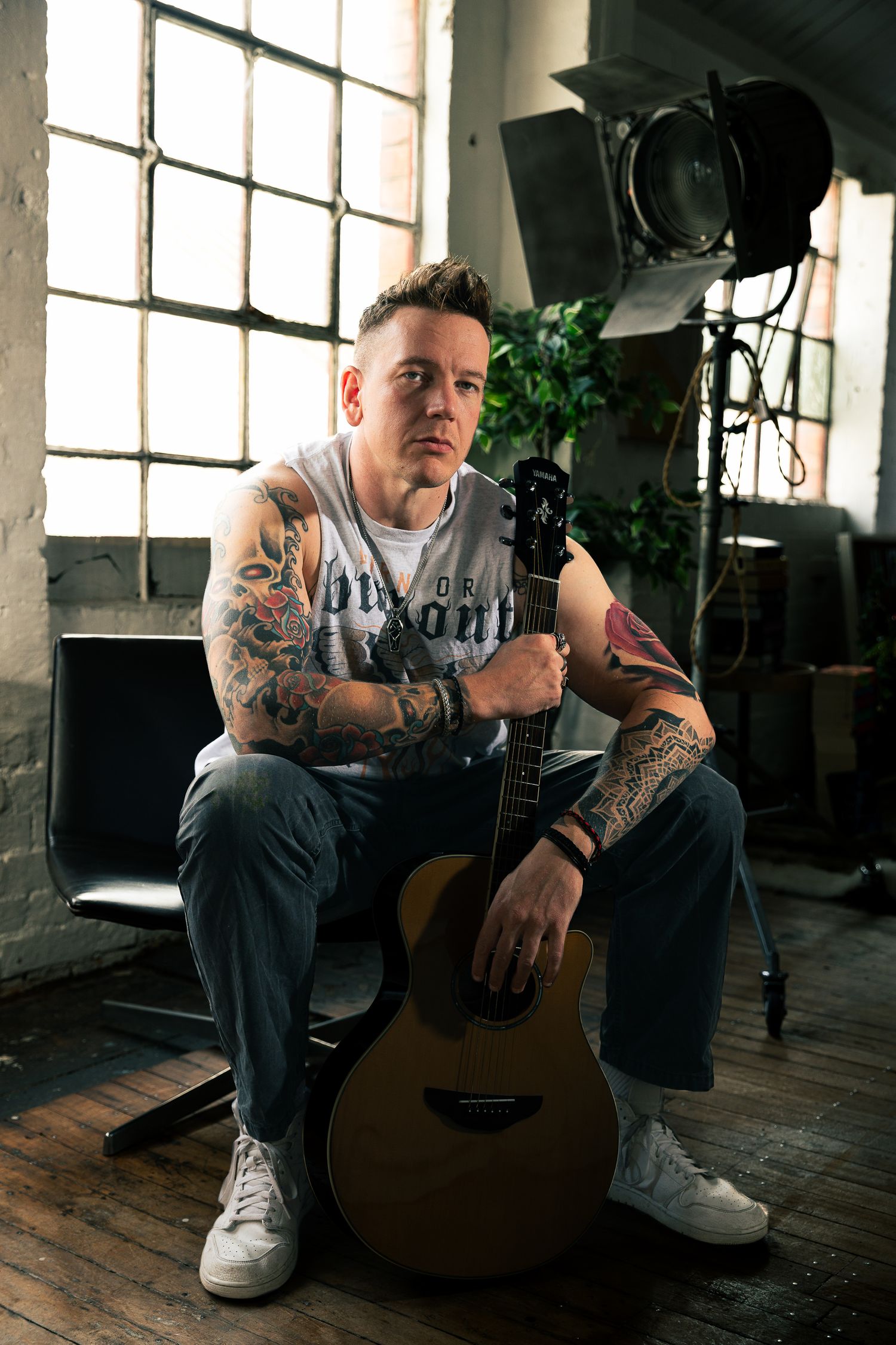 Seated studio portrait of a tattooed man holding an acoustic guitar, soft window light and creative workspace