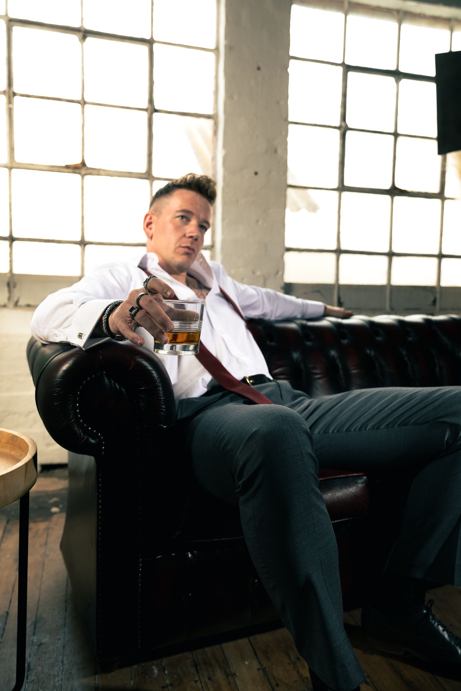 Editorial-style portrait of a man lounging on a leather sofa in a bright loft, holding a drink and looking off-camera.