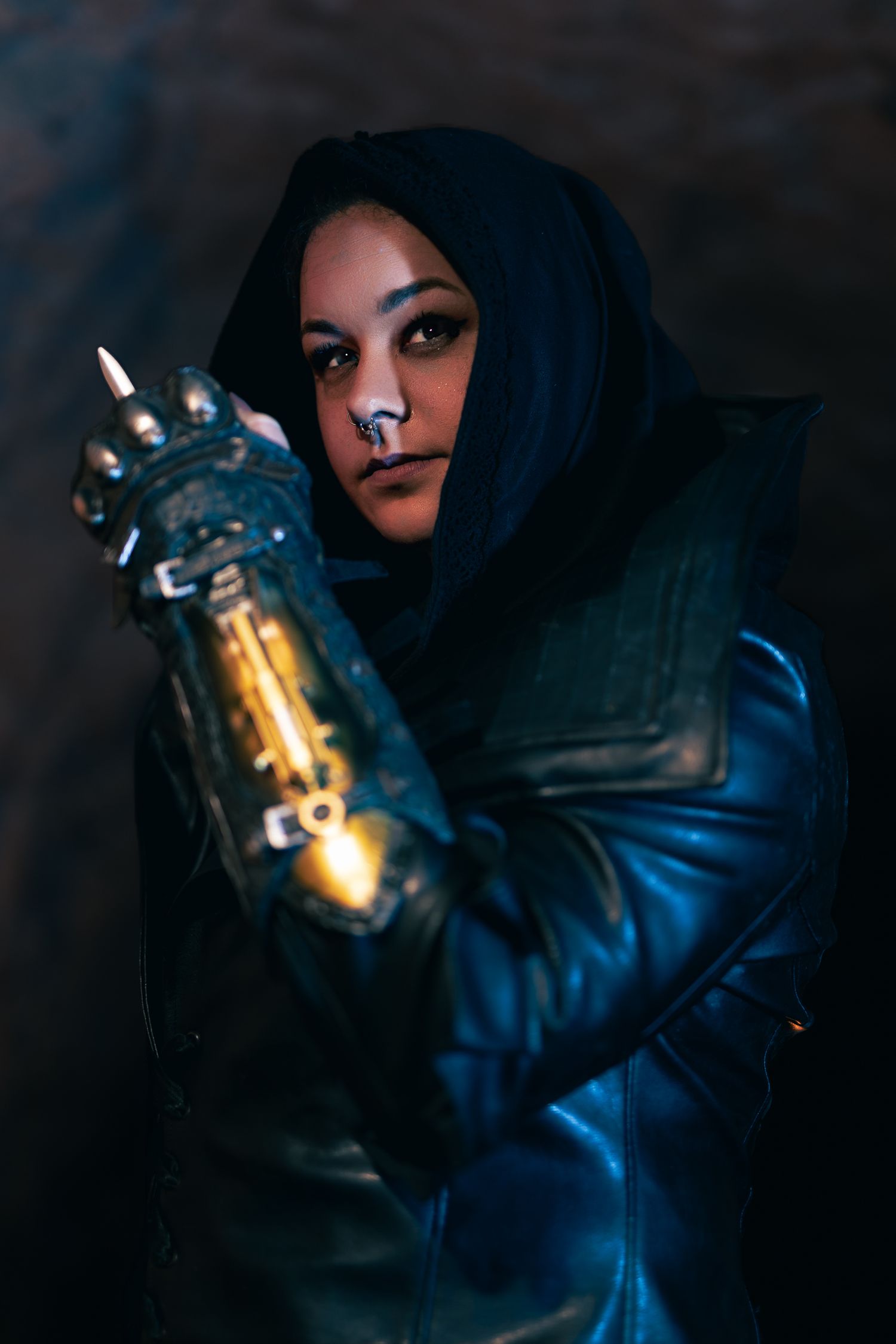 Woman in dark hood and leather jacket holds a glowing mechanical gauntlet in dramatic cosplay photo.