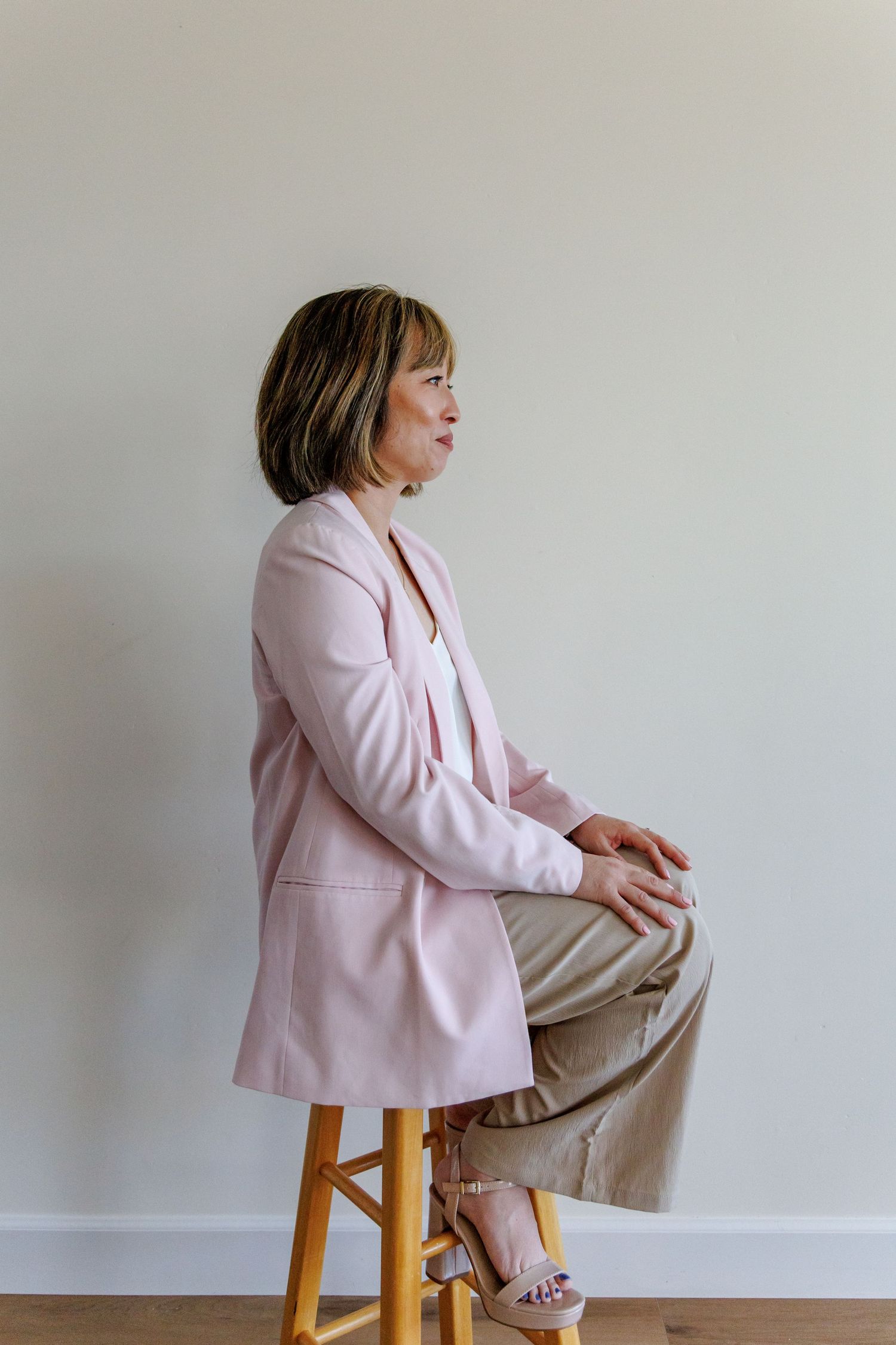 A woman in a pink blazer and beige pants sits on a wooden stool against a light gray wall.