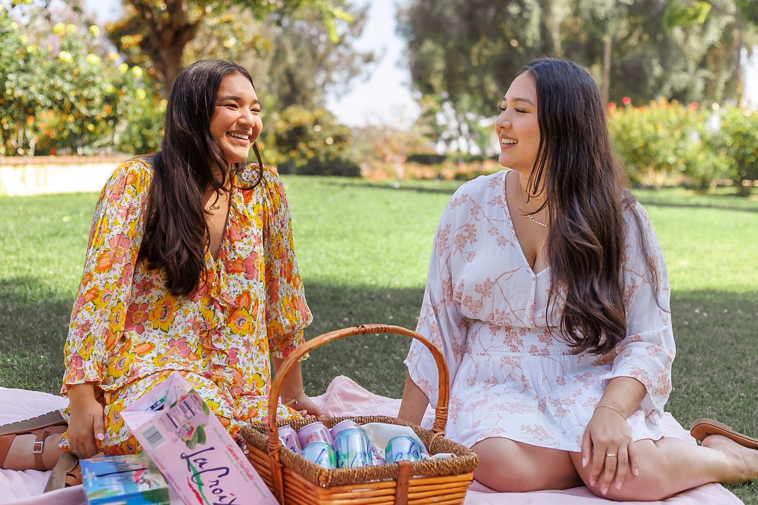 Two smiling women sitting on a blanket outdoors at a picnic with a wicker basket of colorful canned drinks.