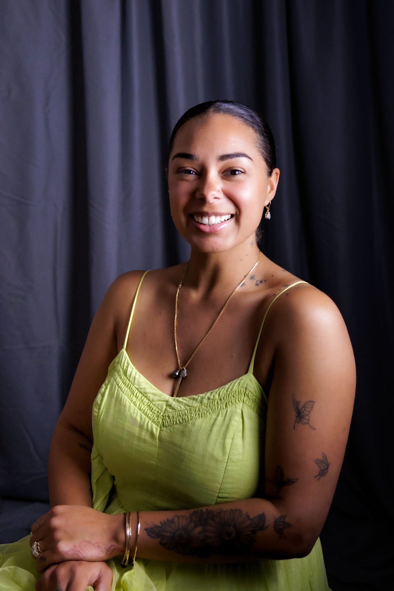 Smiling woman with tattoos wearing a lime green sundress seated against dark gray backdrop.