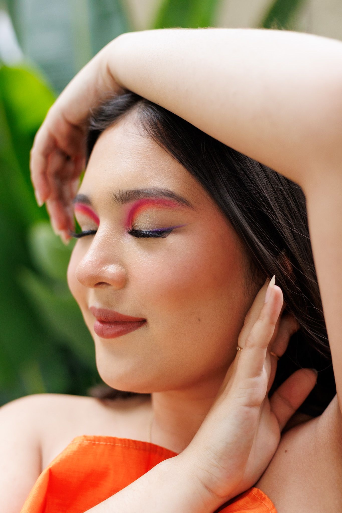 Young woman with vibrant pink eye makeup poses with eyes closed and hands near face outdoors.