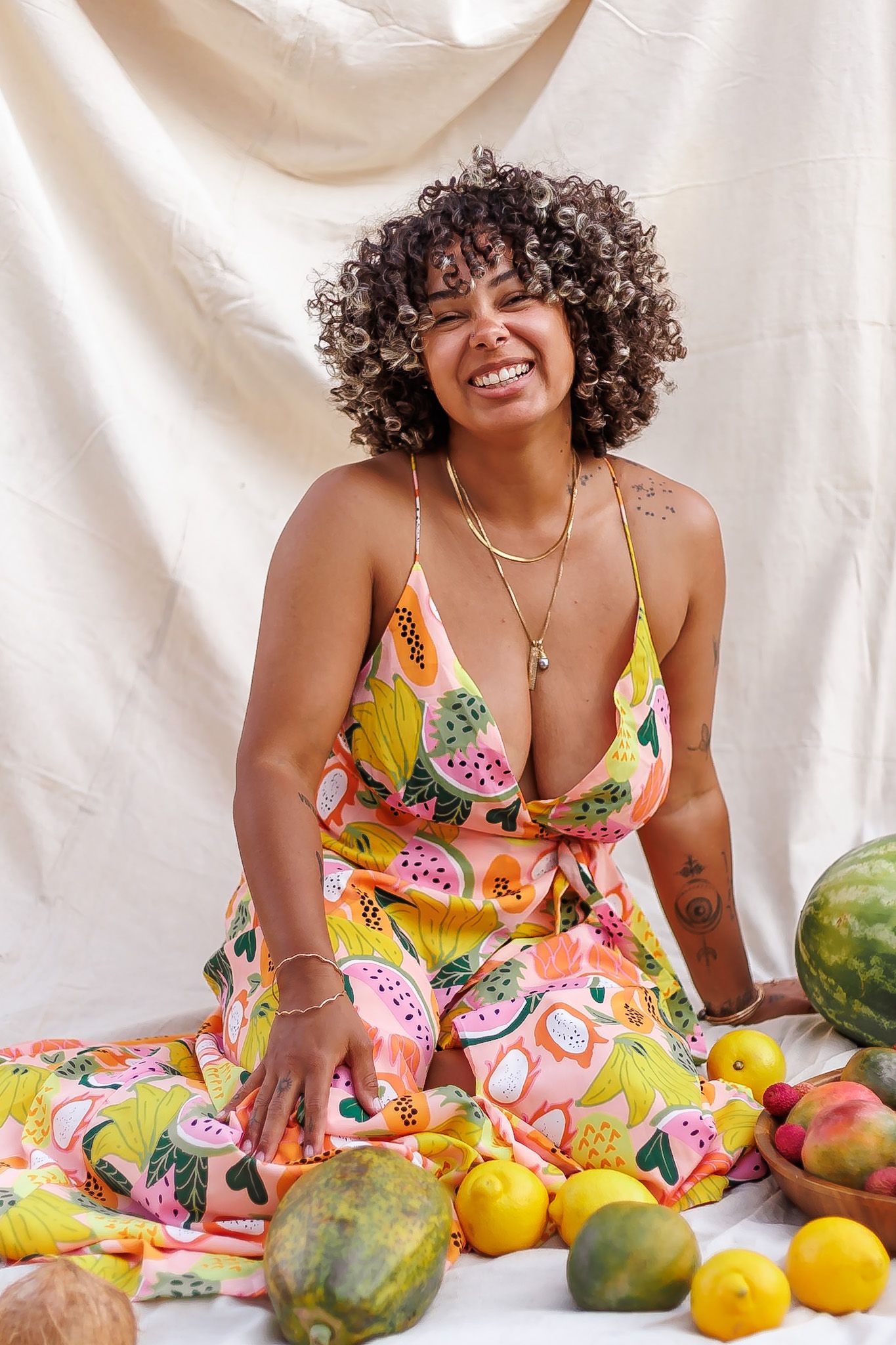 Smiling woman with curly hair sits in colorful fruit-print dress surrounded by tropical fruits.