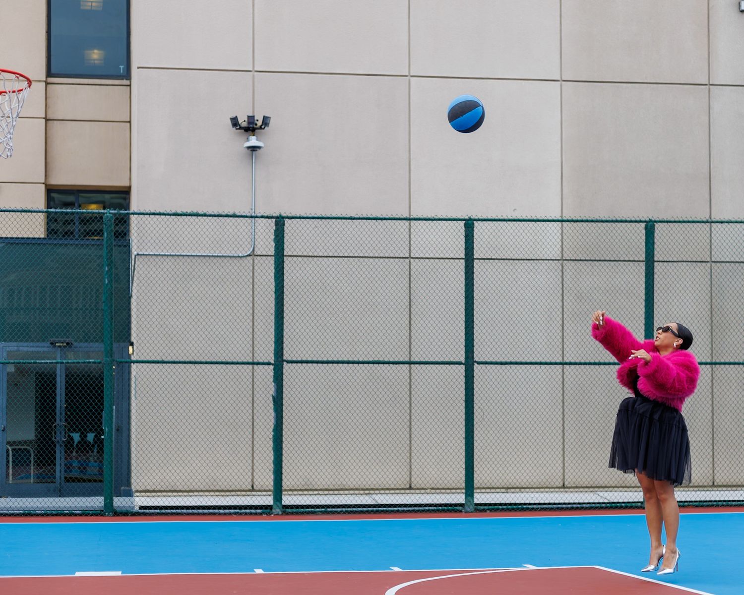Woman in pink fur coat shooting a basketball on an outdoor court with chain-link fence.