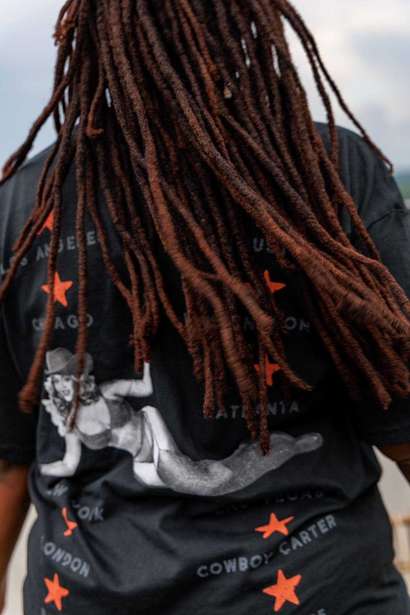 Back view of person with long brown locs wearing black Cowboy Carter graphic t-shirt with orange stars.
