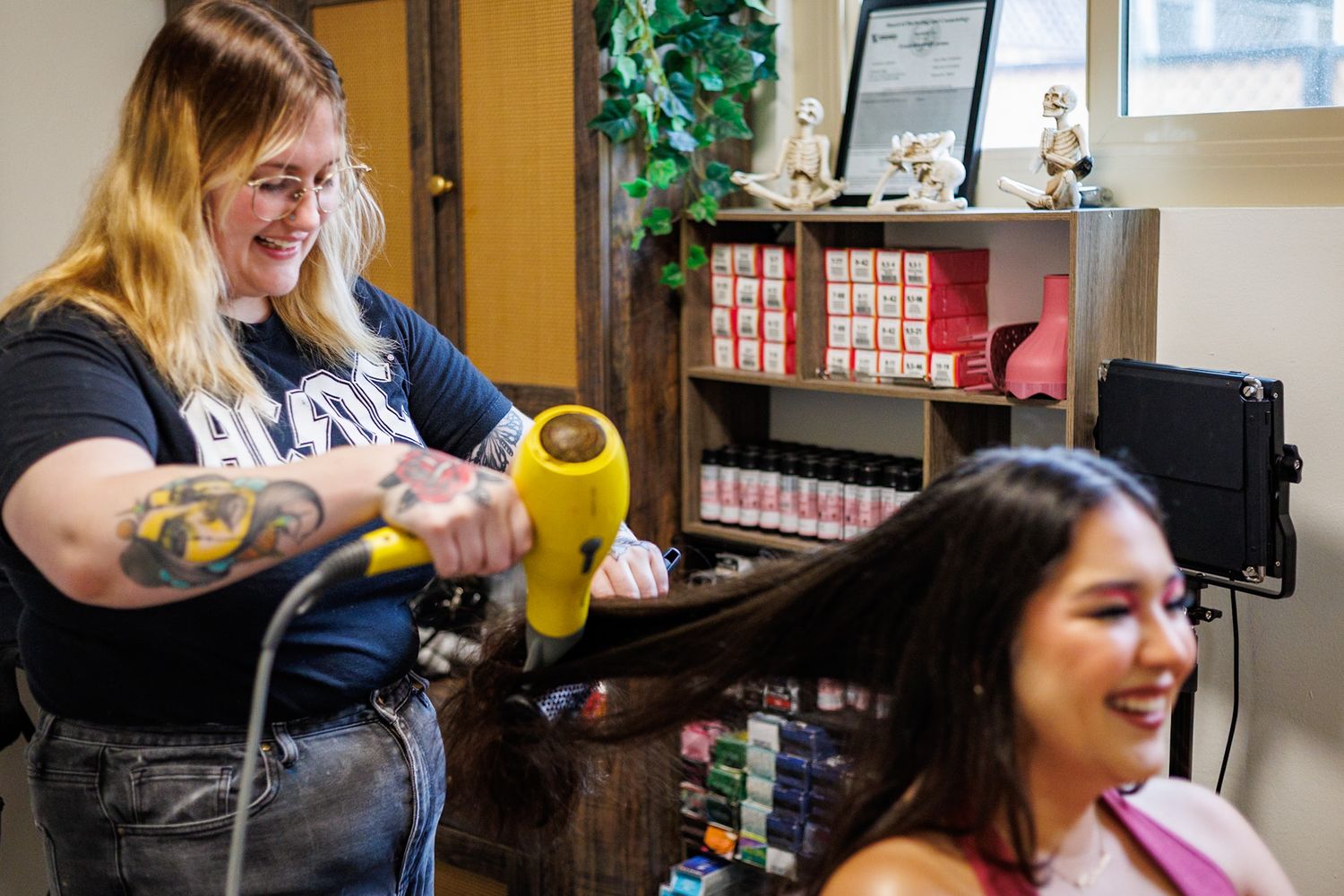Smiling tattooed hairstylist blow dries long dark hair of happy client in bright salon.