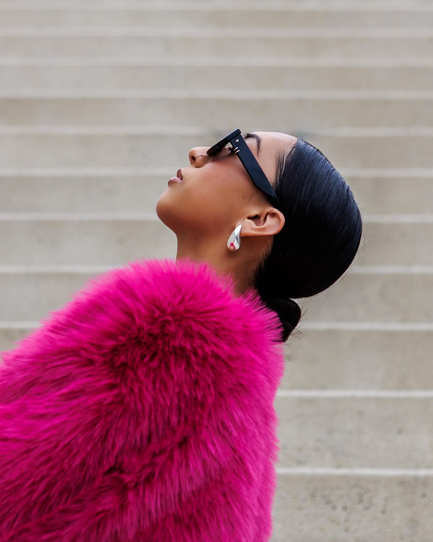 Stylish woman in vibrant pink fur coat wearing black sunglasses and earbuds looking upward.