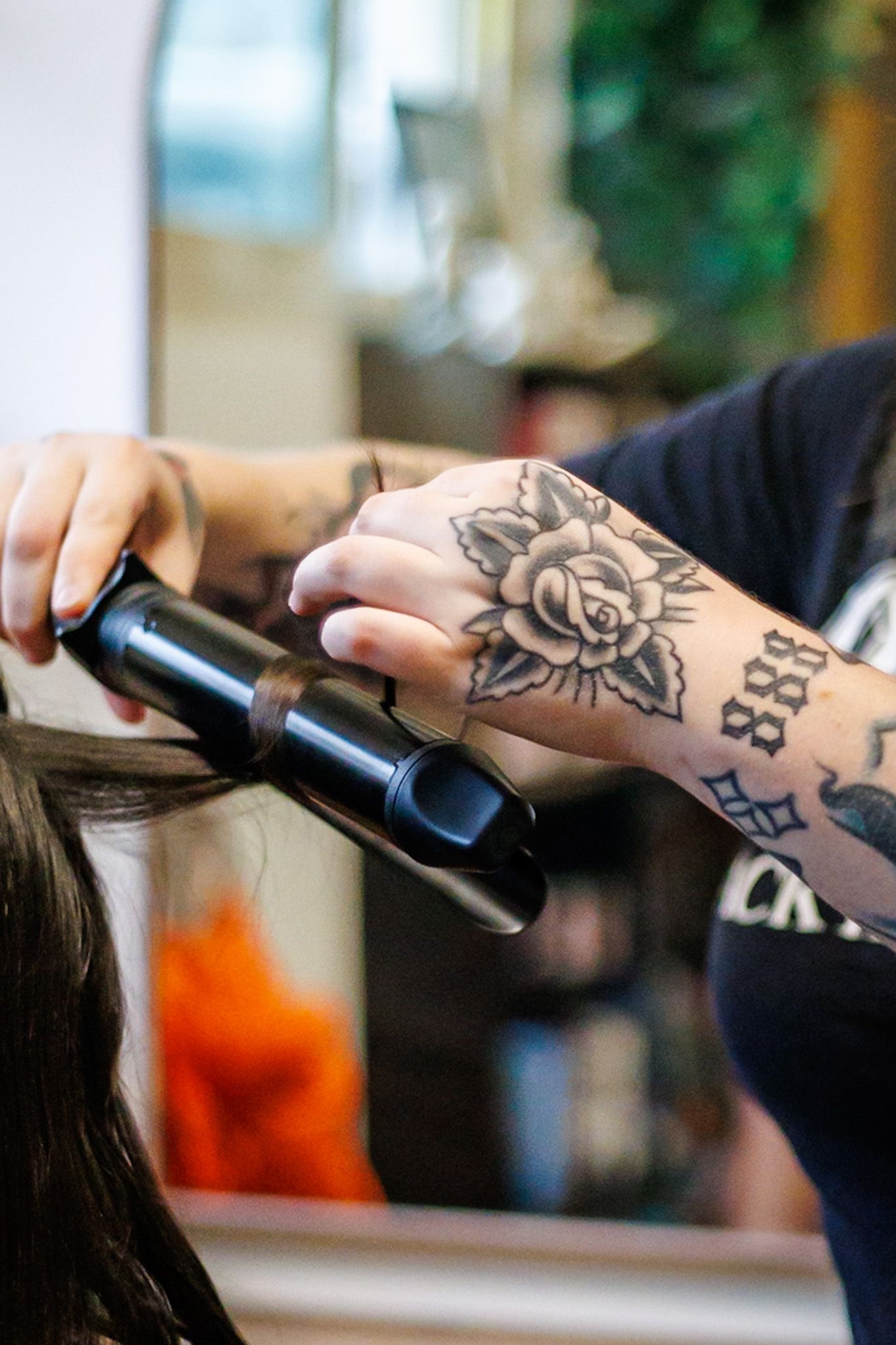 Tattooed hairstylist uses curling iron on client's dark hair in salon with mirror backdrop.