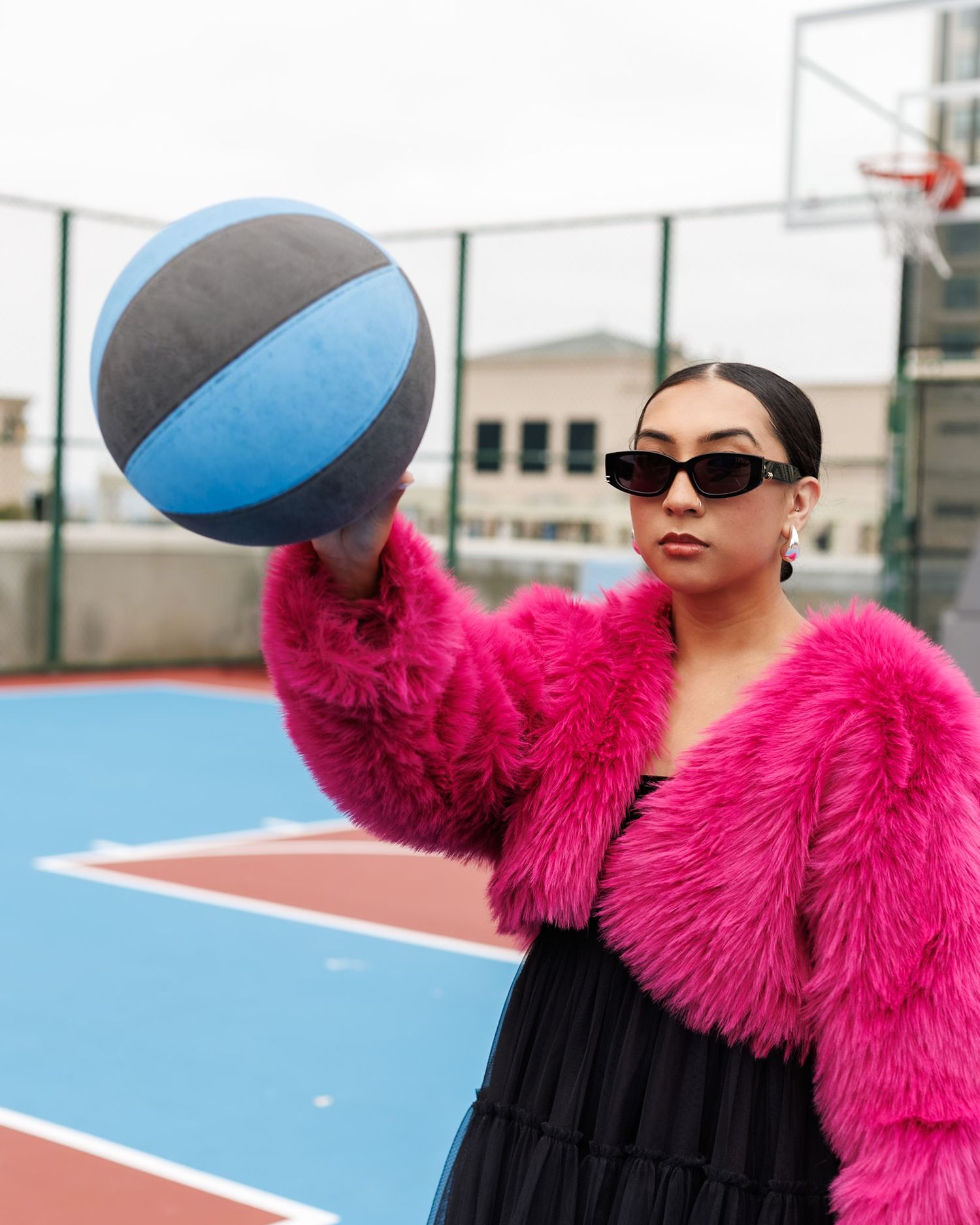 Fashionable woman in hot pink fur coat holding basketball on outdoor court wearing sunglasses.