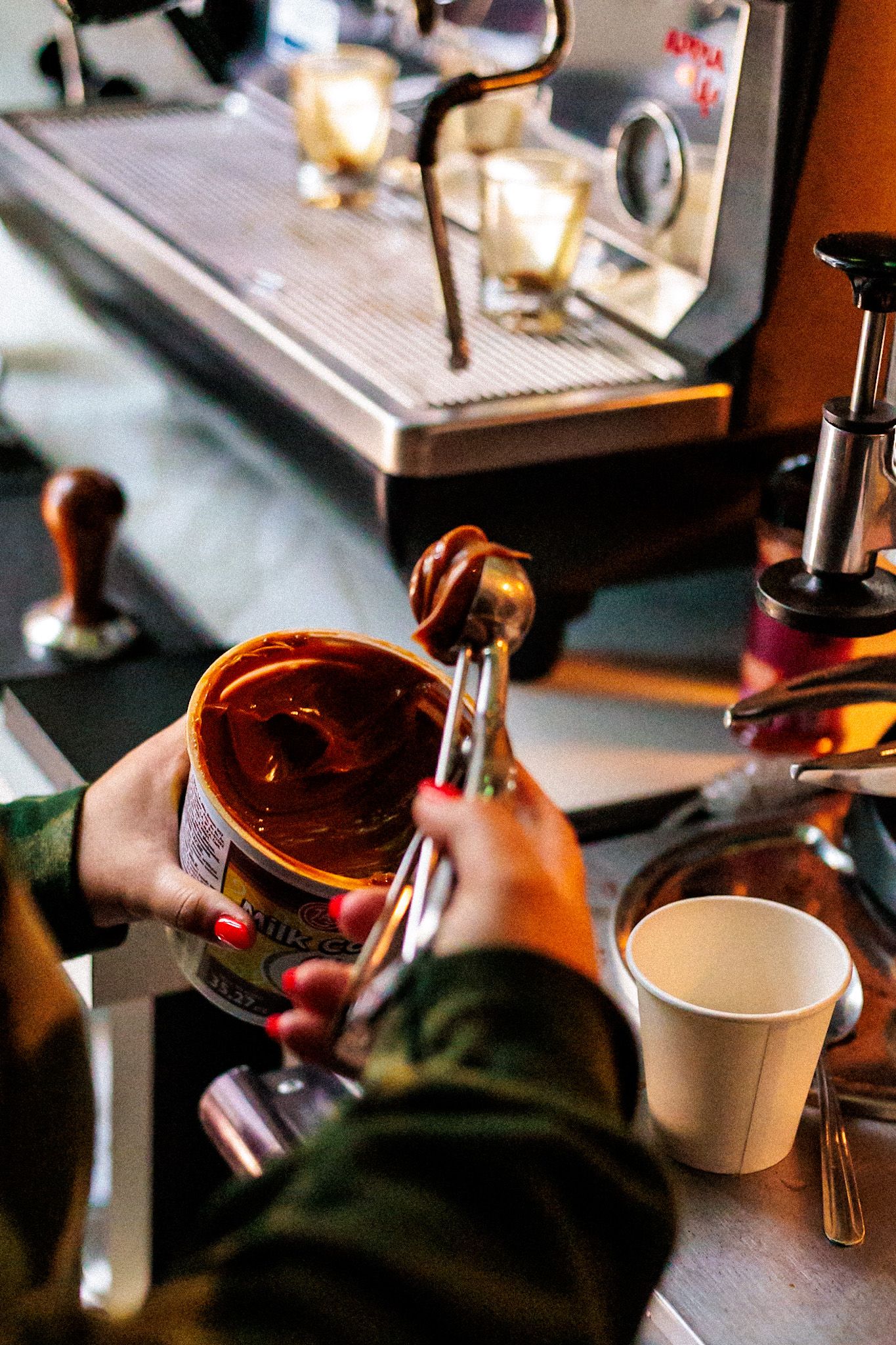 Barista scooping Milo powder with red-nailed hands near an espresso machine at a cafe counter.