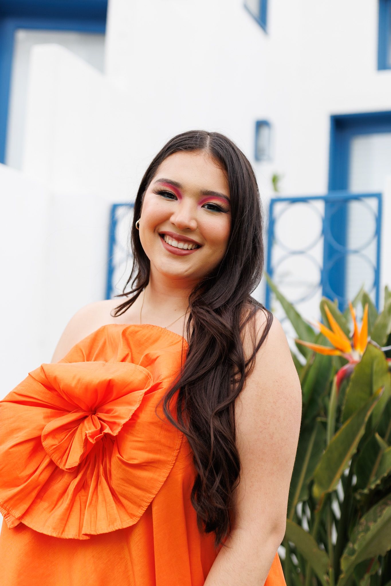 Smiling young woman in vibrant orange dress with floral detail and bold red eye makeup posing outdoors.