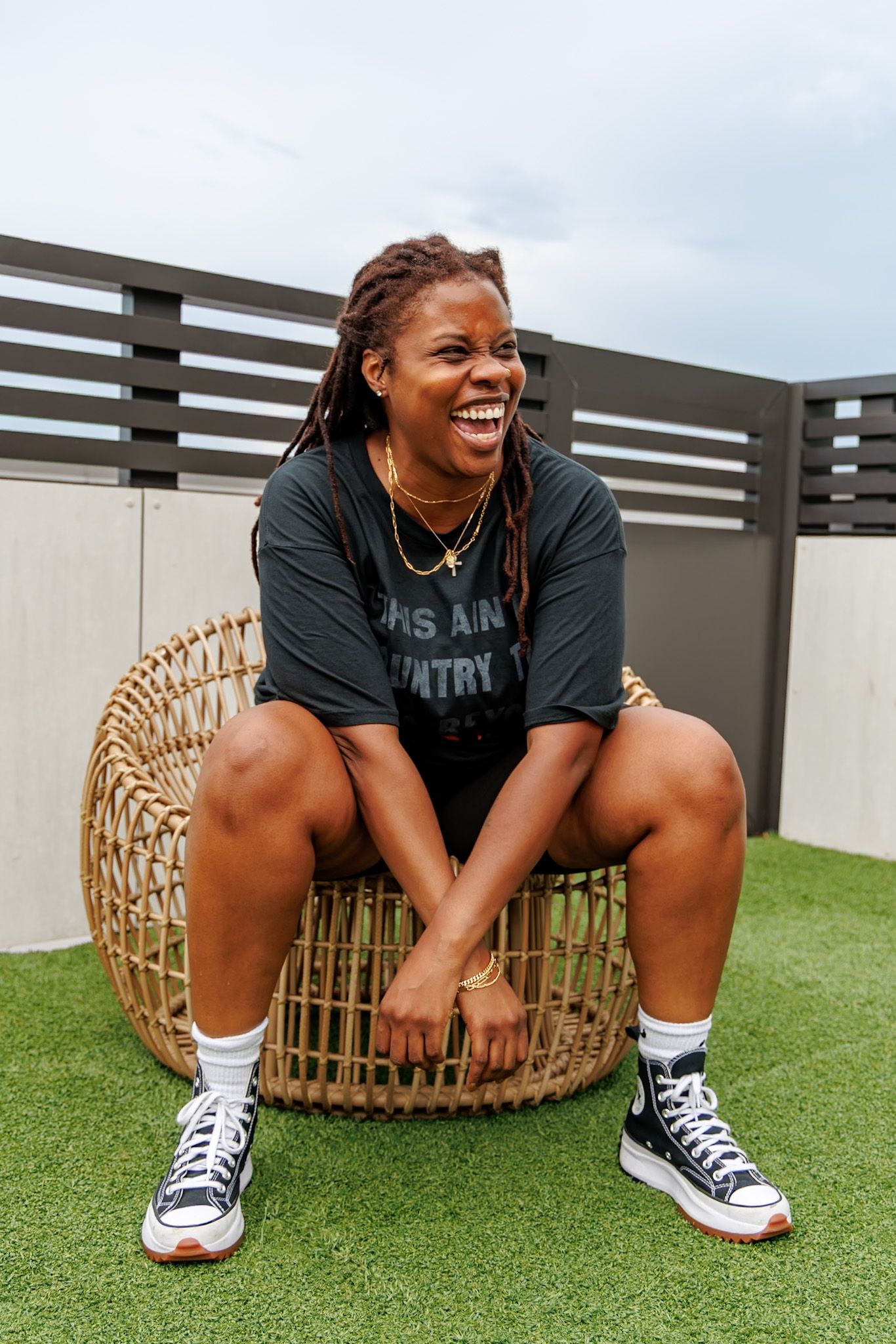 Laughing Black woman with locs in black graphic tee sitting in wicker chair outdoors wearing Converse sneakers.
