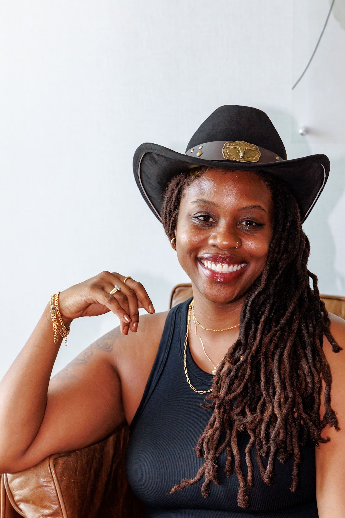 Laughing Black woman with locs wearing black cowboy hat with gold buckle and black tank top on leather chair.