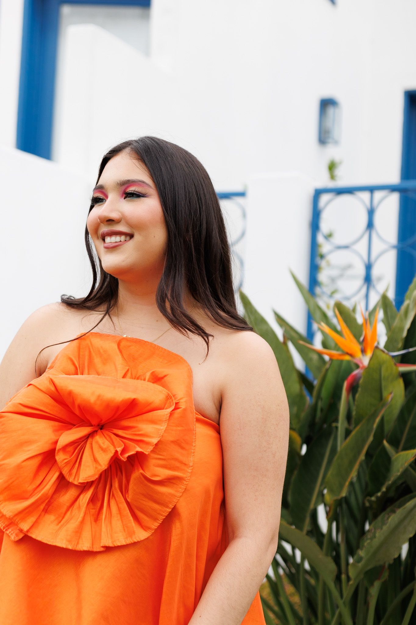 Smiling young woman in orange strapless dress with large flower detail stands near tropical plants.