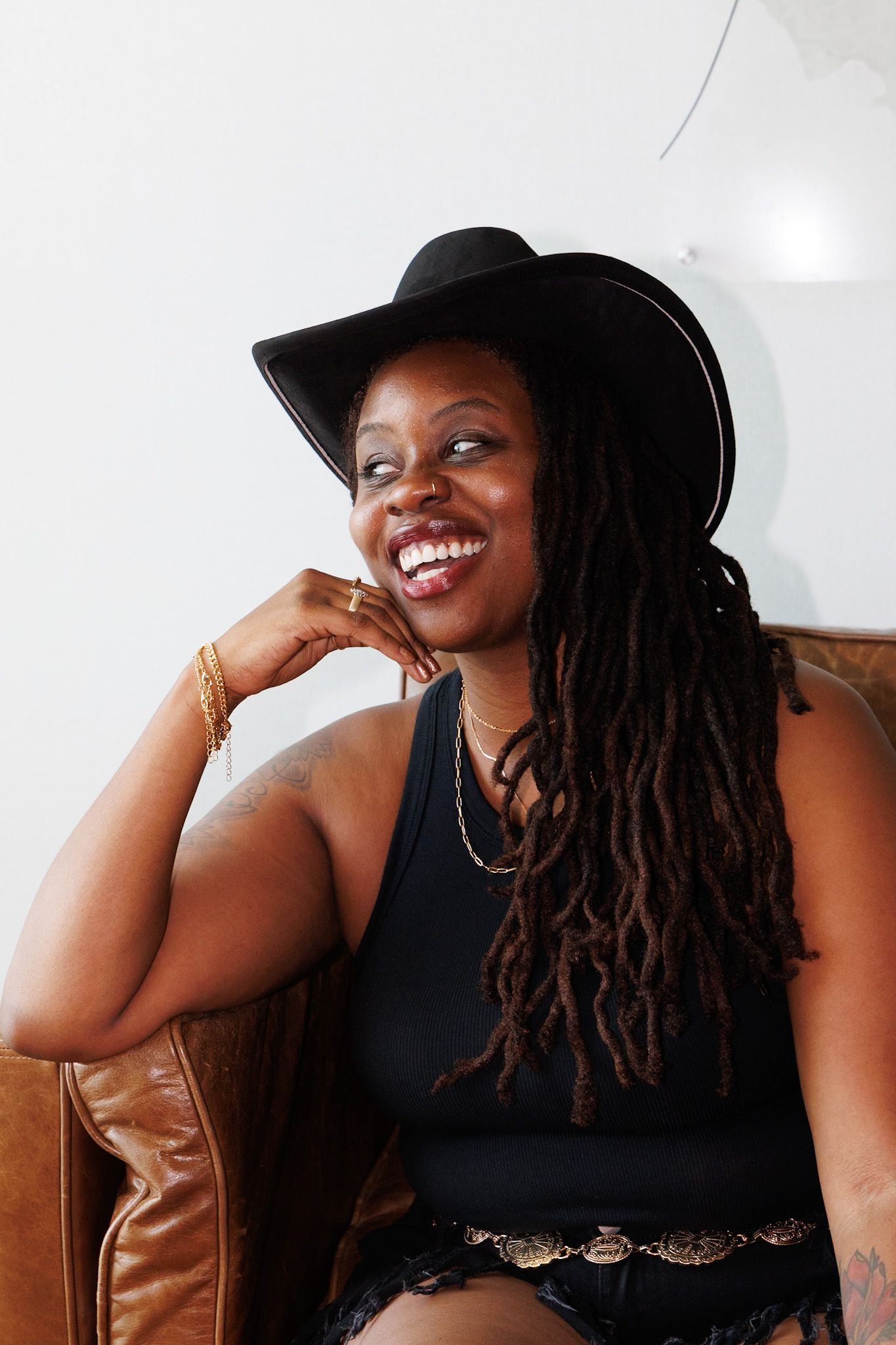 Smiling Black woman with long locs wearing black cowboy hat and black outfit seated on brown leather chair.
