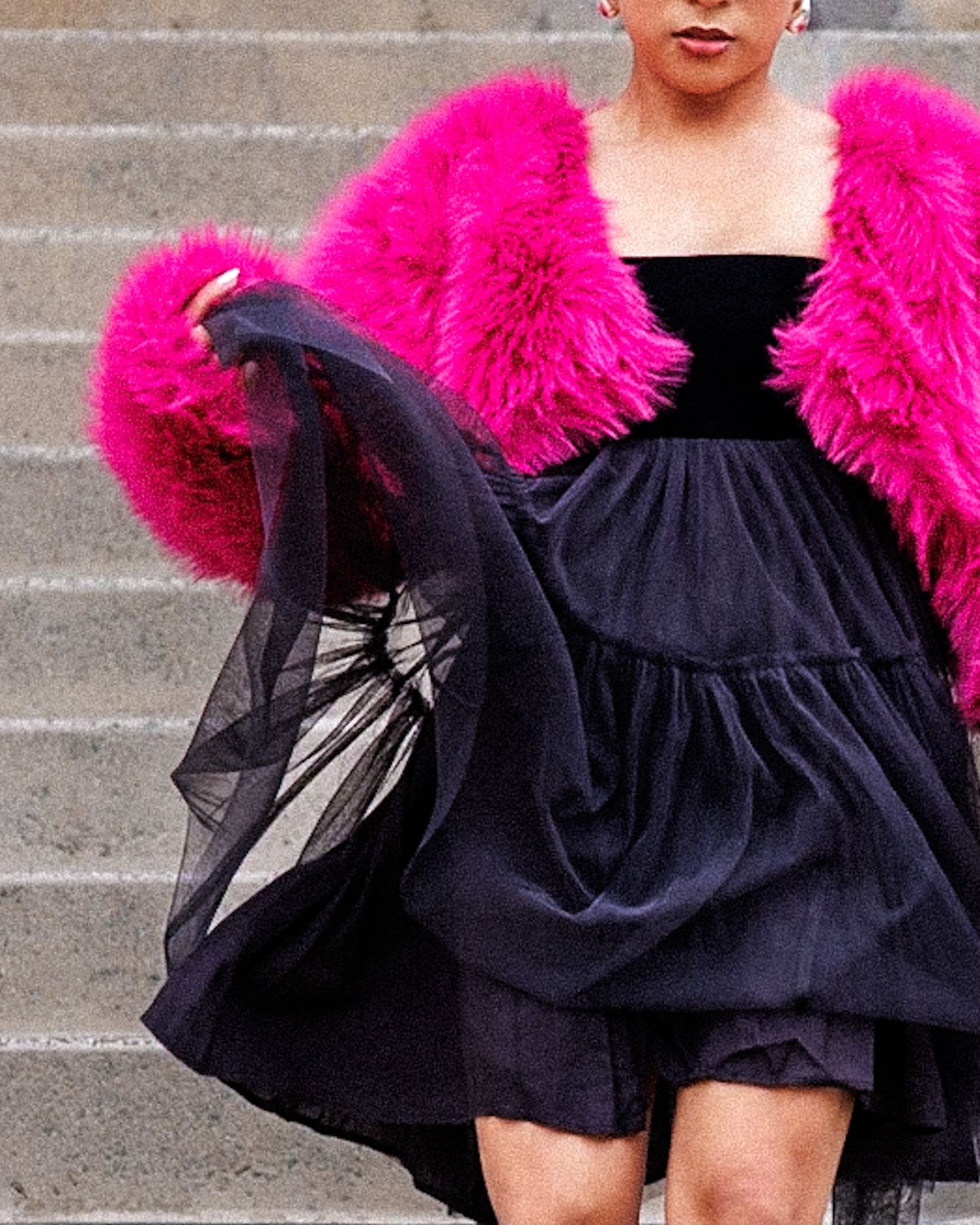 Close-up of woman wearing hot pink fur coat over black strapless tiered dress on steps.