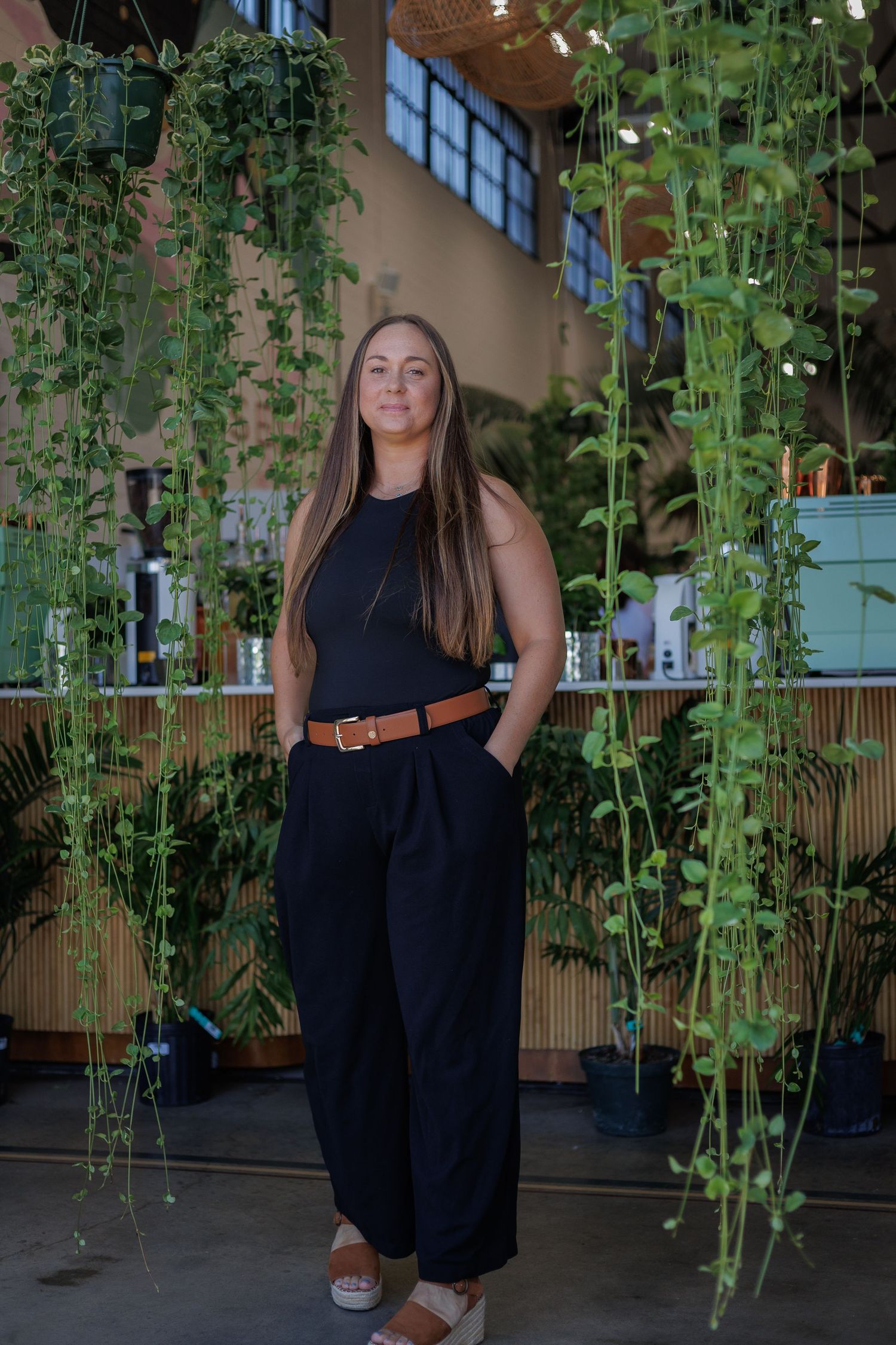 A woman in a black sleeveless outfit stands among hanging green plants in a stylish indoor space.