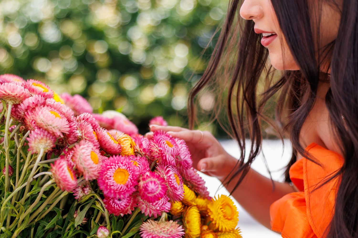 Young woman in orange dress smelling vibrant pink and yellow everlasting flowers outdoors.