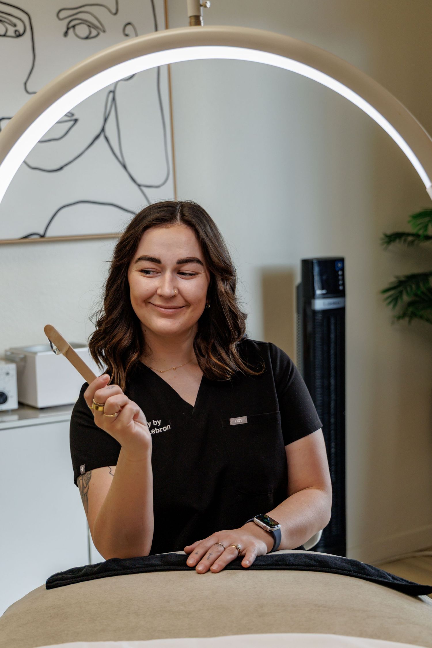 A smiling esthetician in black uniform holds a nail file under a ring light at a beauty salon.
