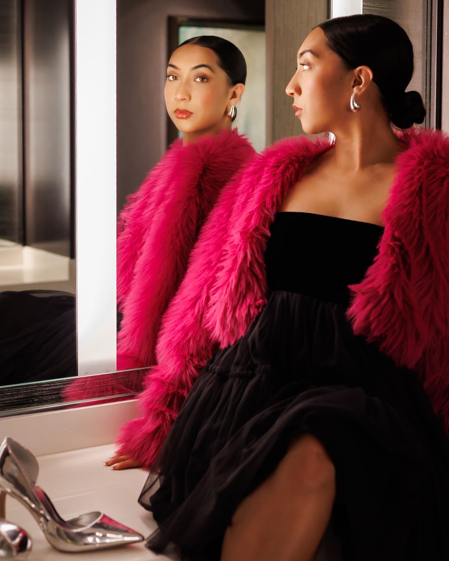 Elegant woman in hot pink faux fur coat and black dress reflected in mirror with silver heels nearby.
