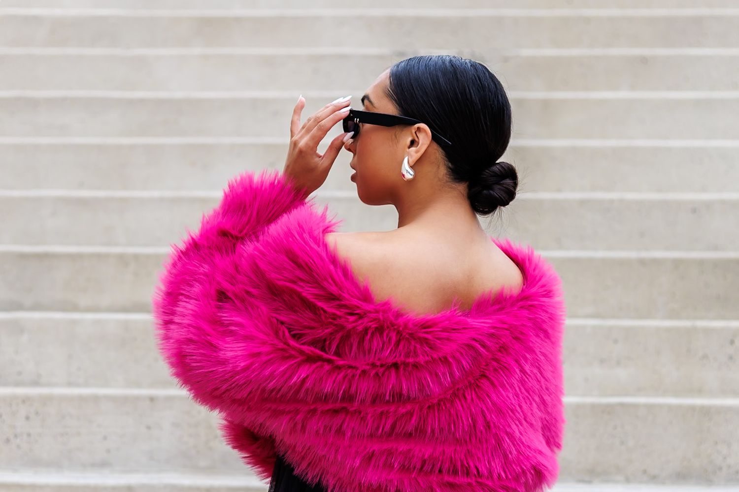 Woman in hot pink off-shoulder fur coat wearing sunglasses shielding eyes on outdoor steps.