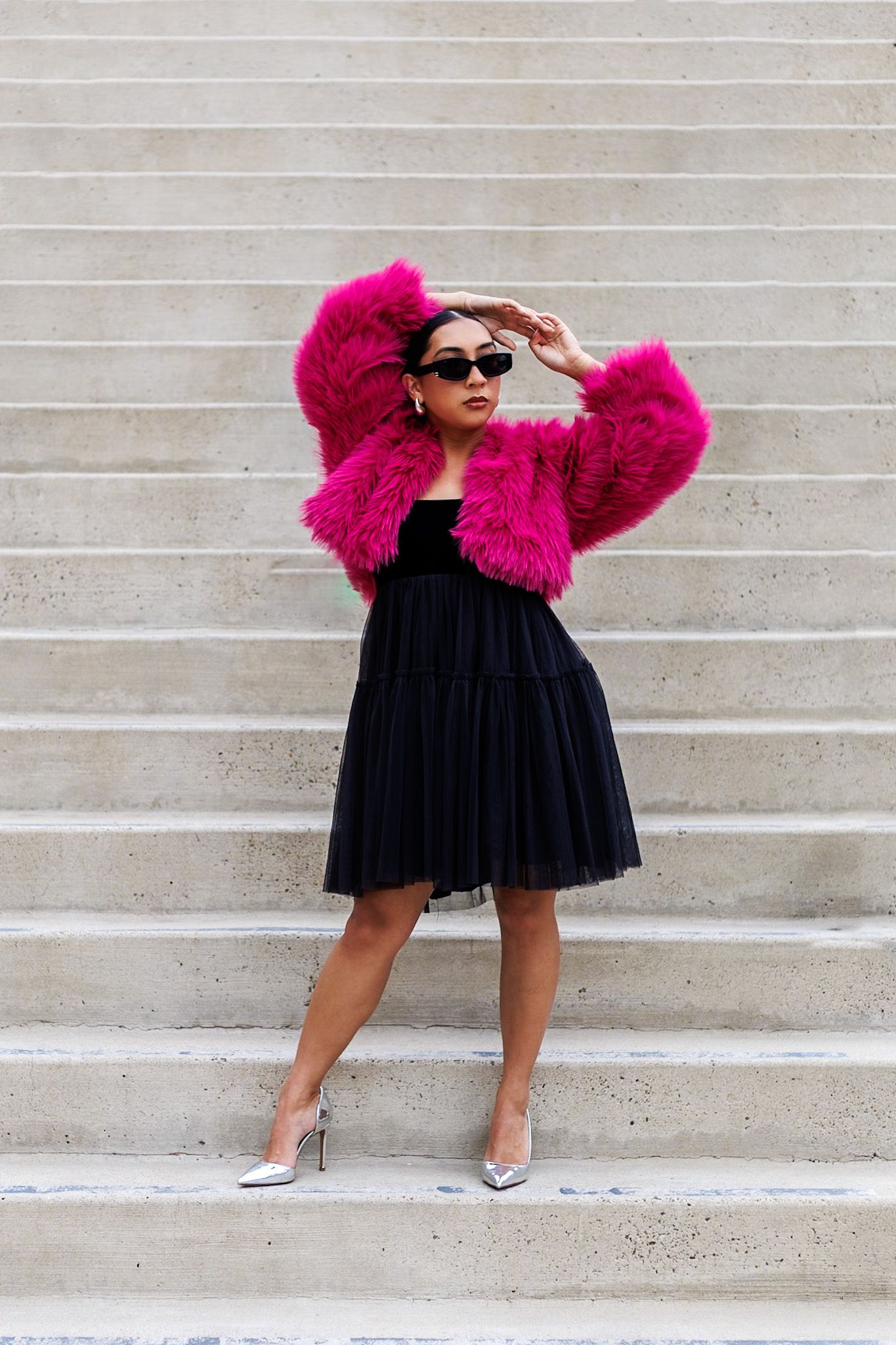 Confident woman in hot pink fur coat and black tulle skirt posing on stone steps.