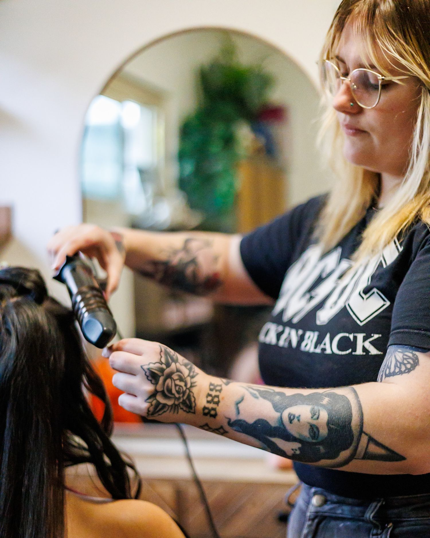 Tattooed female hairstylist in AC/DC shirt styles long dark hair with curling iron in salon.