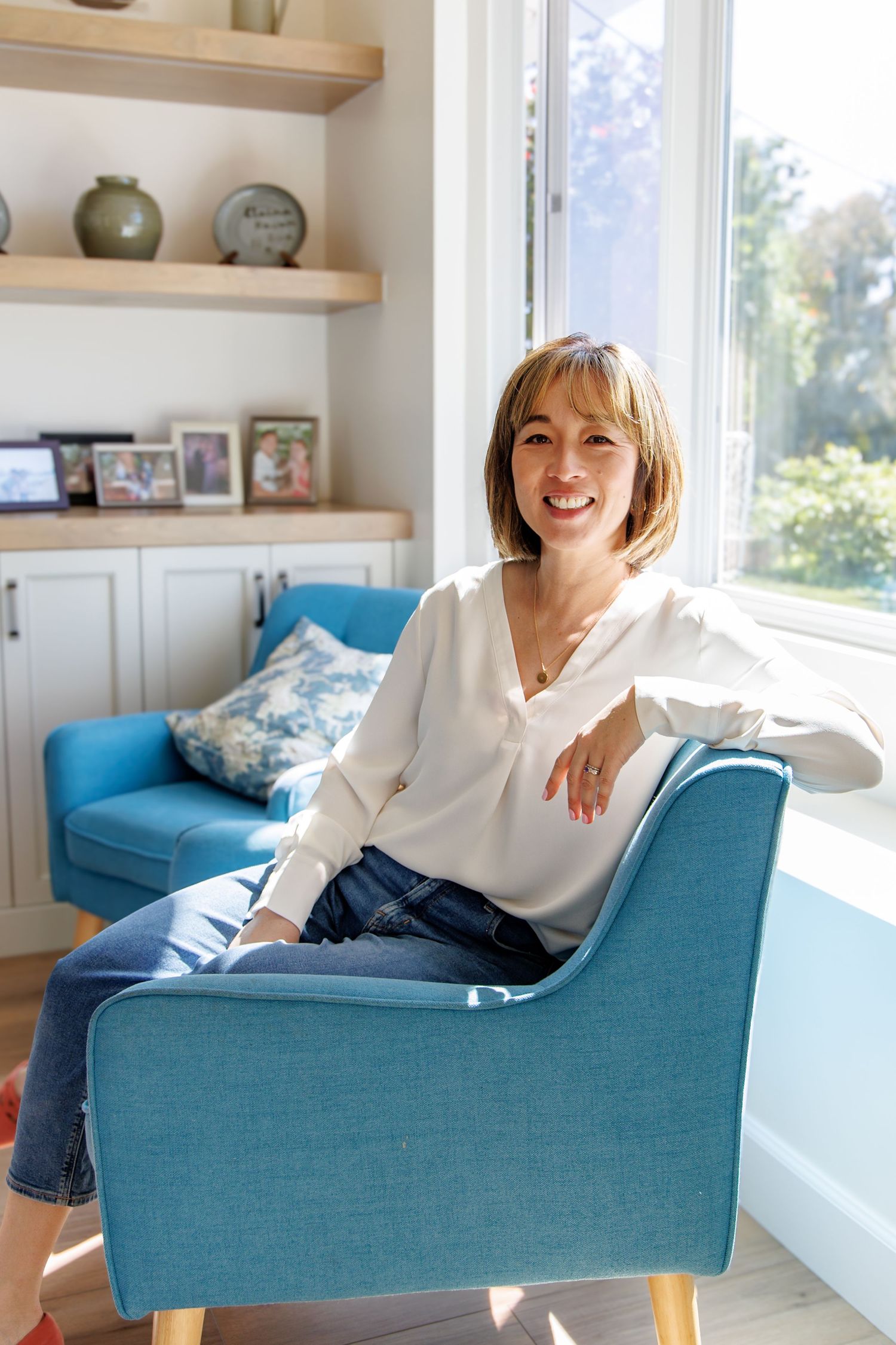 Smiling woman with bob haircut sitting in blue chair by sunny window in bright modern home.