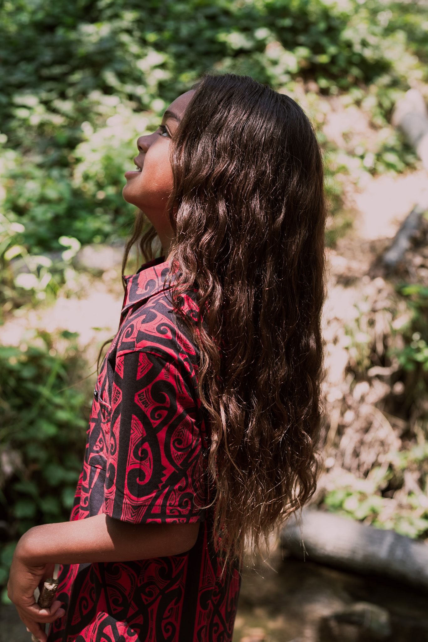 Side profile of a boy with long wavy hair wearing a red and black tribal print shirt outdoors.