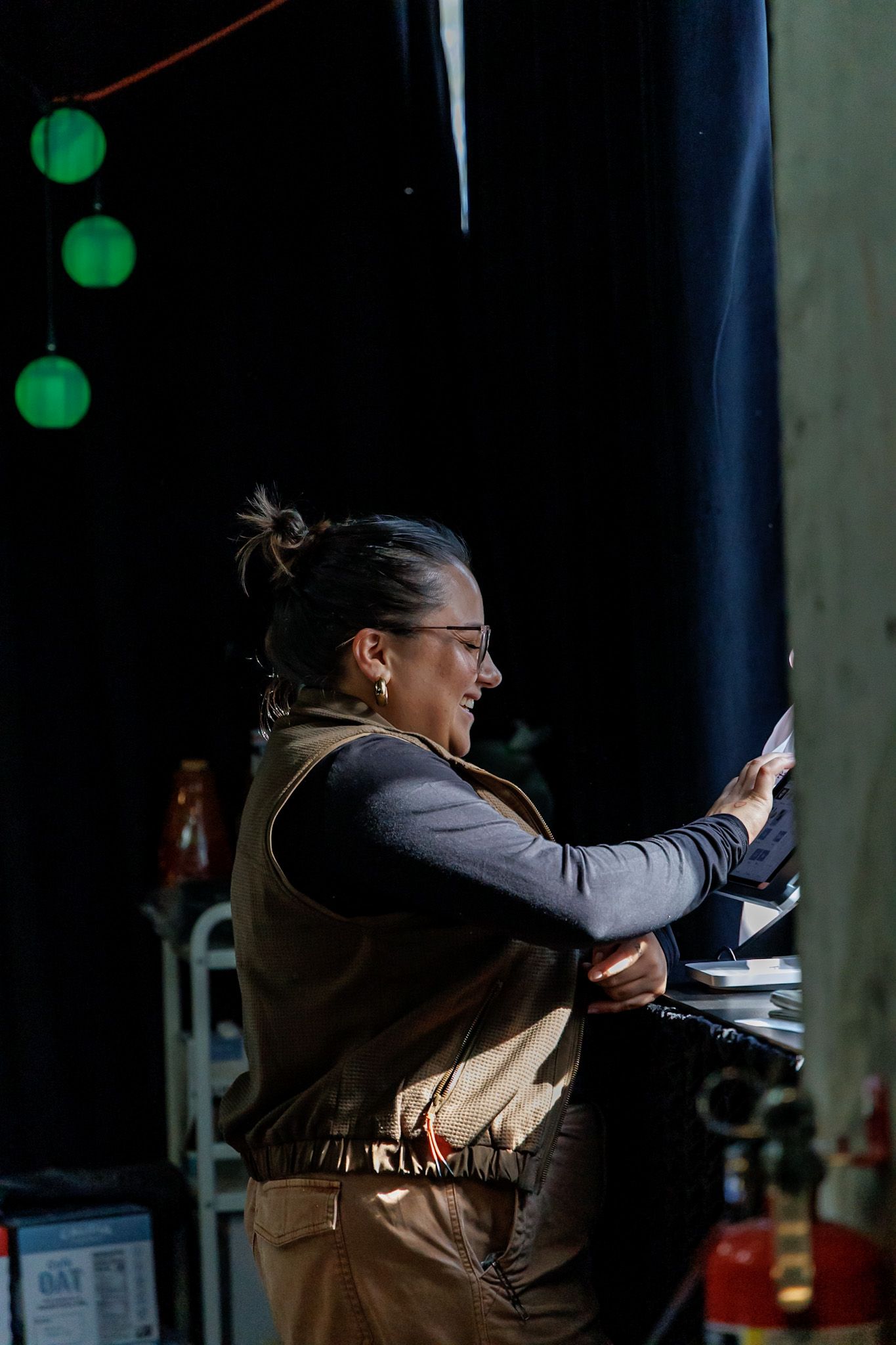 A smiling woman in glasses and a brown vest works at a dimly lit cafe counter under green pendant lights.