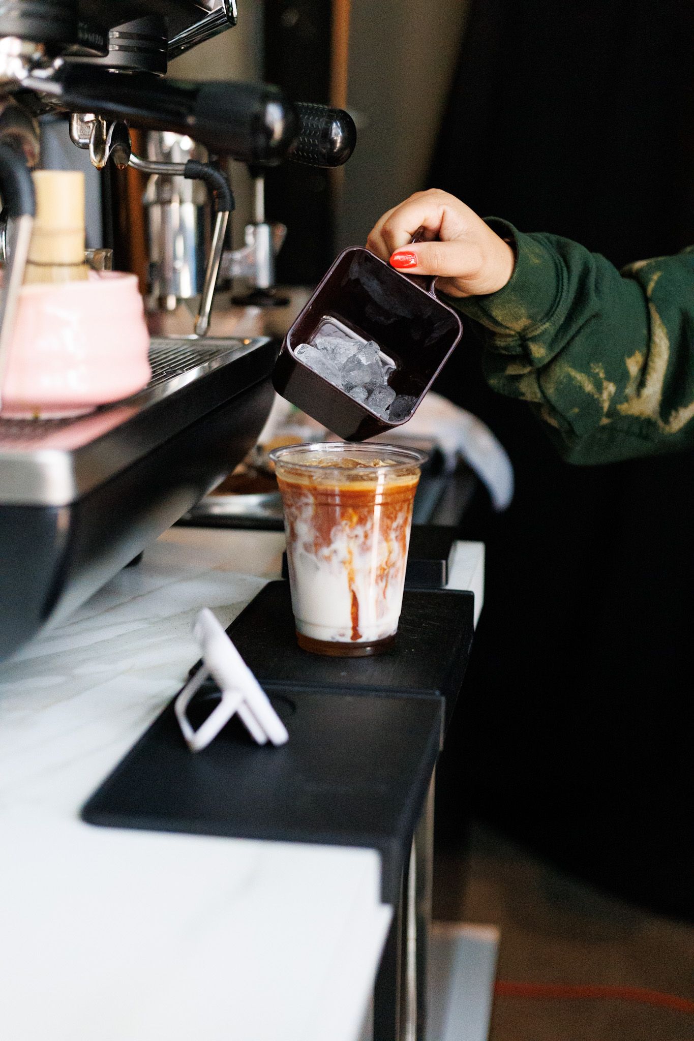 Barista pouring ice into an iced latte in a clear plastic cup at a coffee bar.