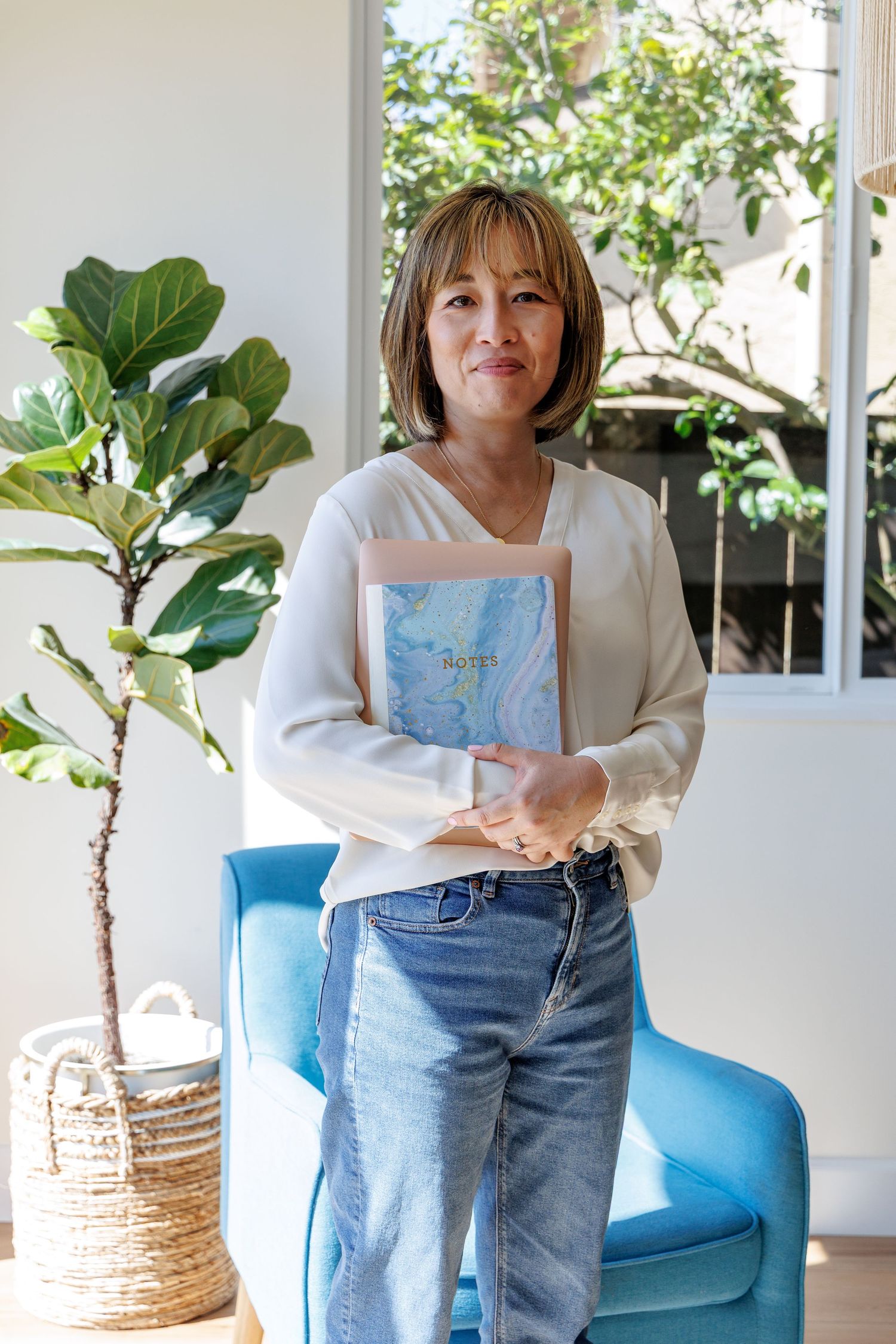 Woman in white blouse and jeans holding a book while standing near a fiddle leaf fig plant.