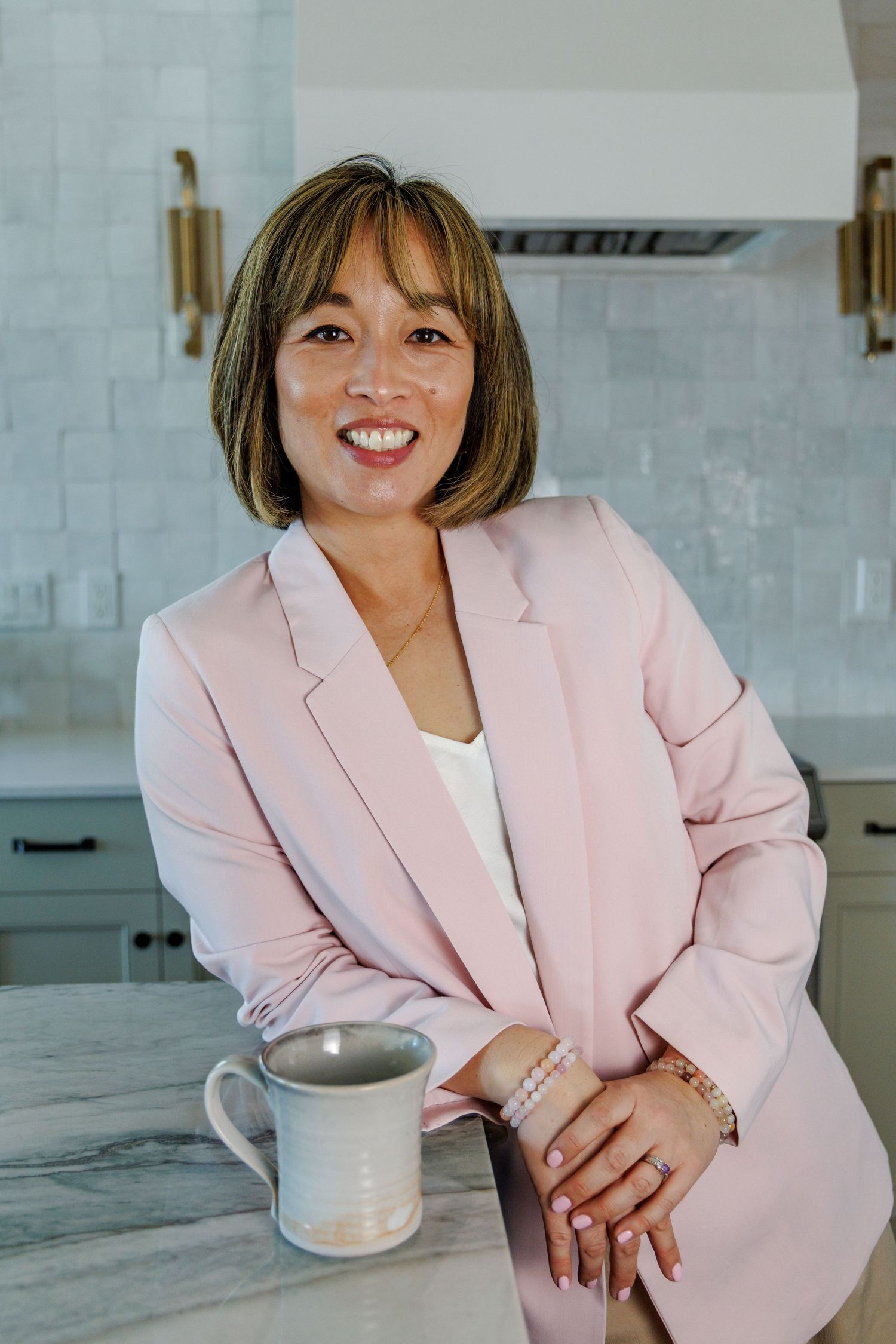 Cheerful woman in pink blazer leaning on marble counter holding white mug in modern kitchen.