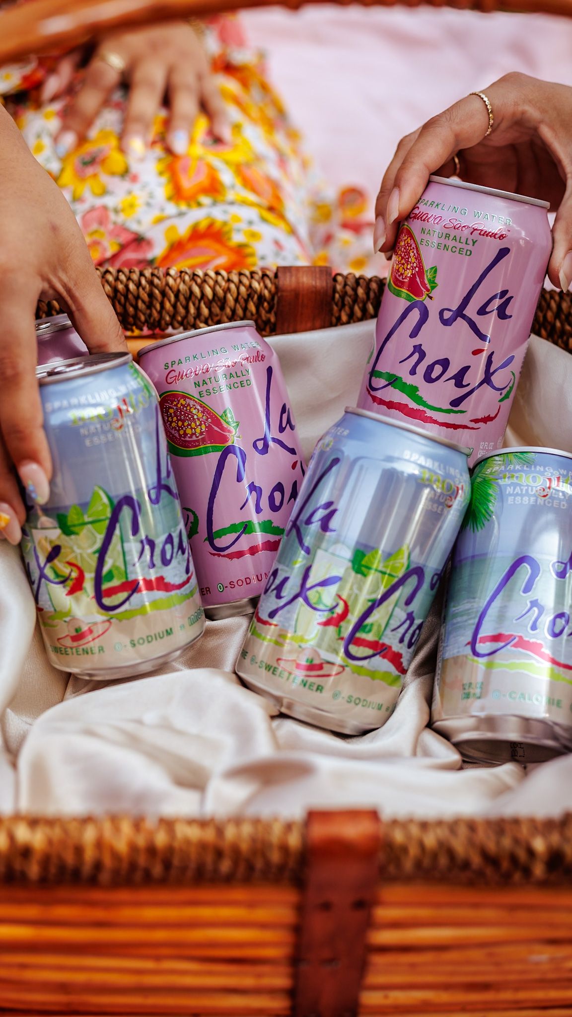 Hands reaching for colorful LaCroix sparkling water cans in a wicker picnic basket lined with white cloth.