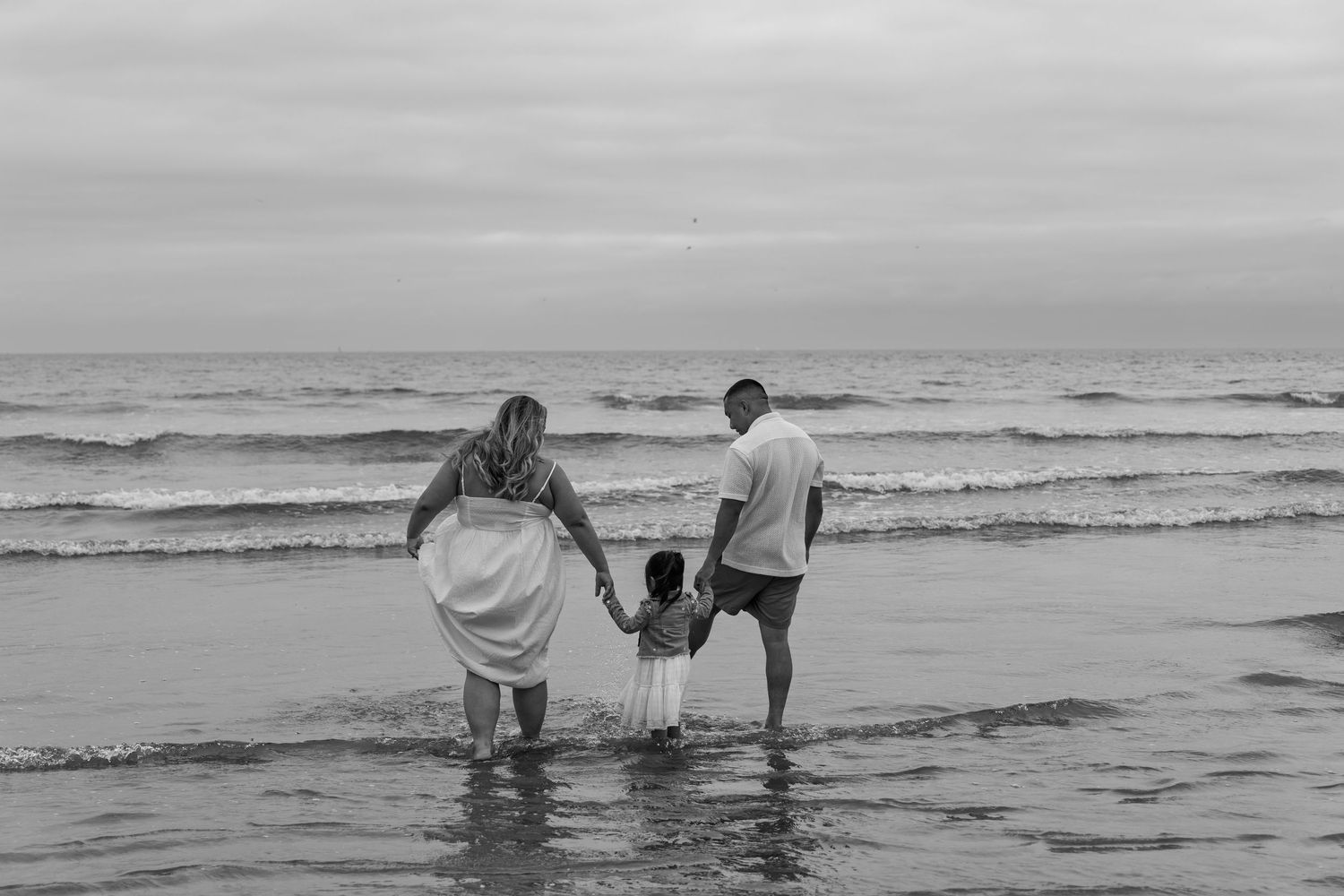 Family of three holding hands walking toward ocean waves on overcast beach in black and white.