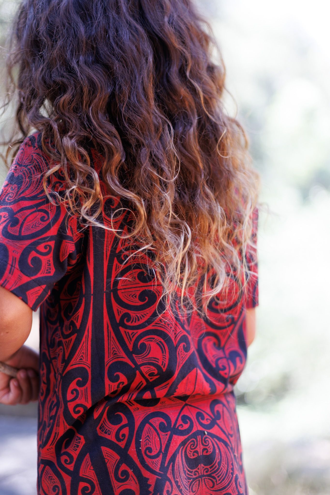 Back view of a boy with curly ombre hair wearing a bold red and black Maori-inspired tribal print shirt.