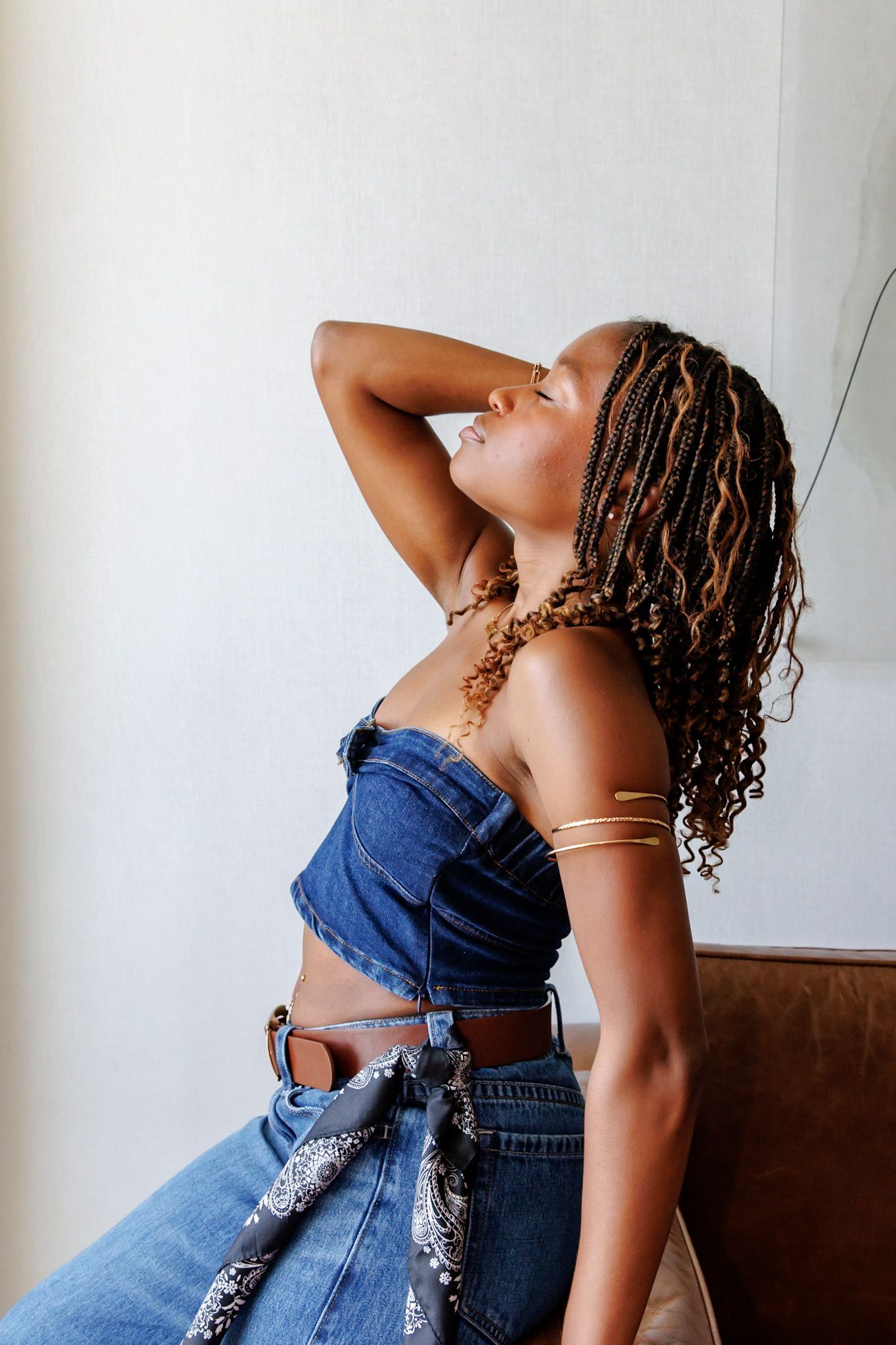 Black woman in denim corset top with gold arm cuff leaning back with hand behind head, eyes closed.