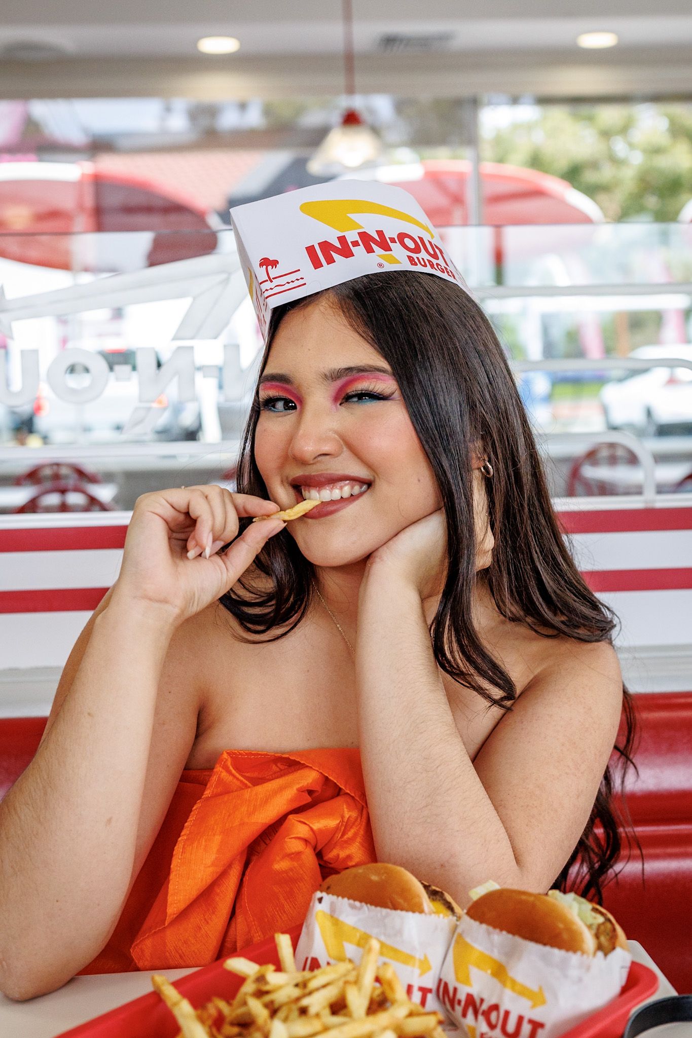 Smiling woman wearing an In-N-Out paper hat eating french fries at the restaurant.
