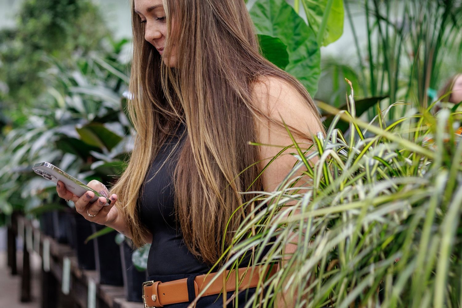 A young woman in a black top and brown belt browses her phone among lush green plants indoors.