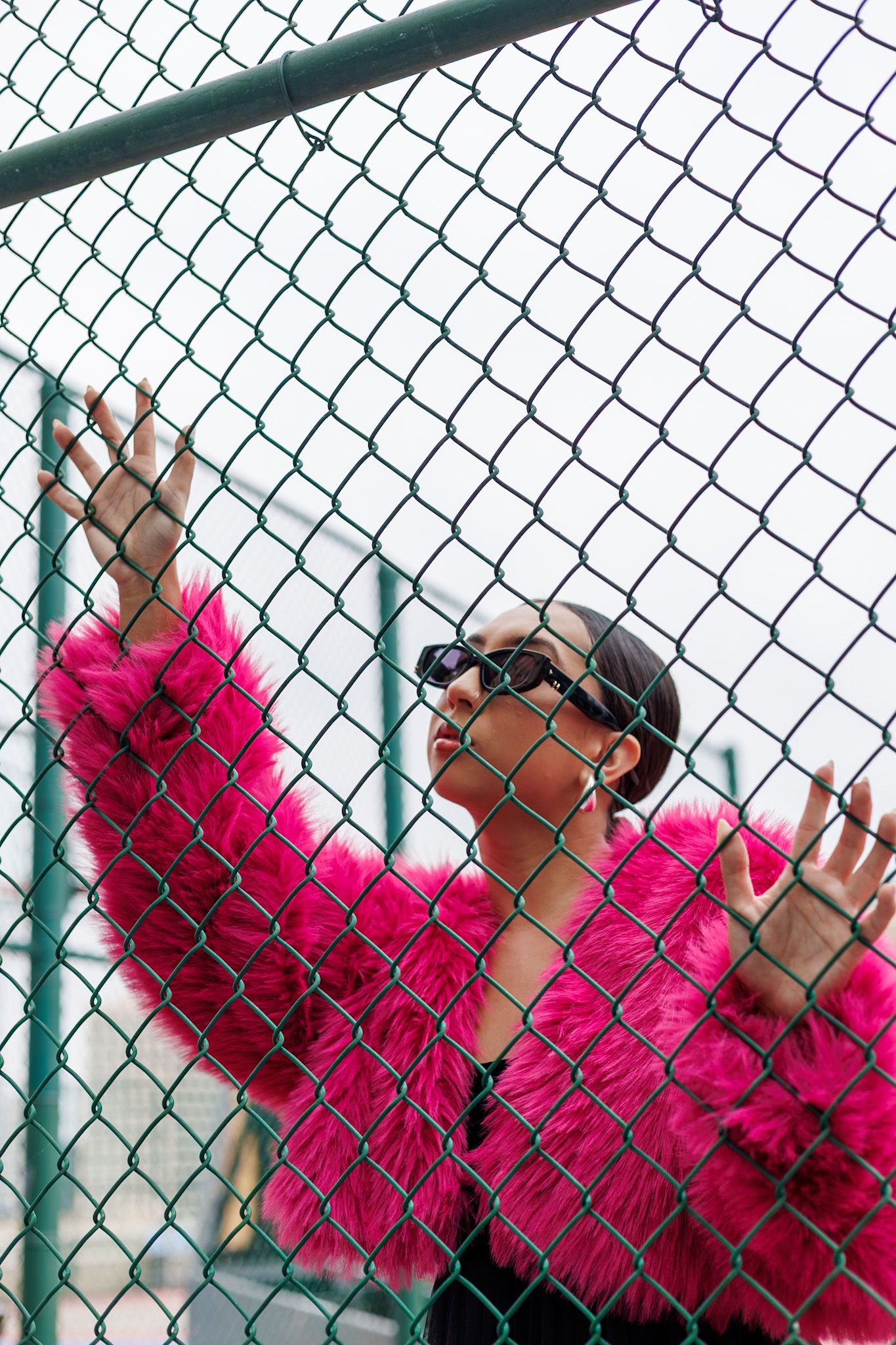 Woman in hot pink faux fur coat and sunglasses standing behind green chain-link fence outdoors.