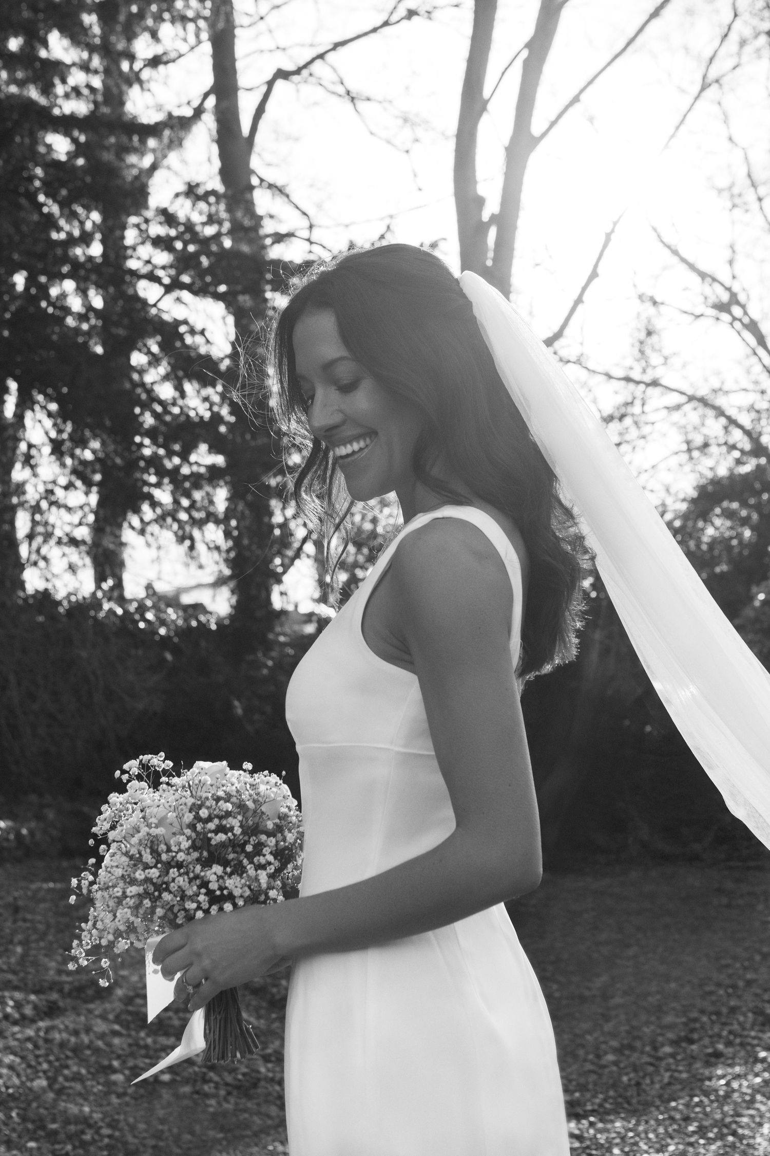 black and white photo of bride smiling, backlit with trees behind