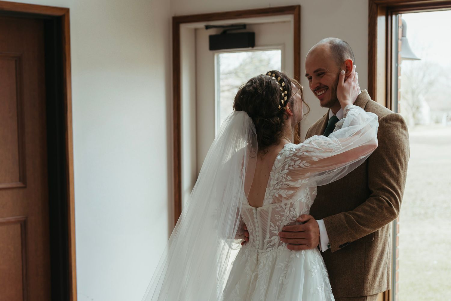 Bride in lace gown and veil sharing first look moment with groom in brown suit, both smiling joyfully.