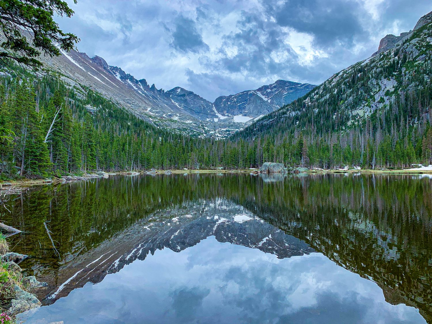 Mountain lake reflecting jagged peaks and pine forest in Rocky Mountain National Park under dramatic summer clouds.
