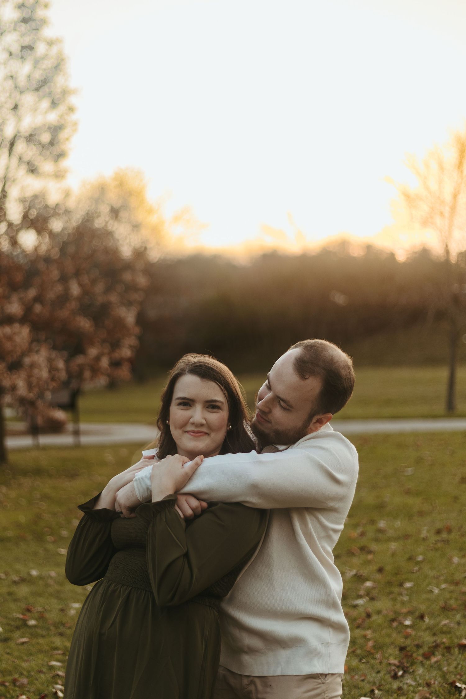 Man embracing woman from behind smiling at camera in autumn park.